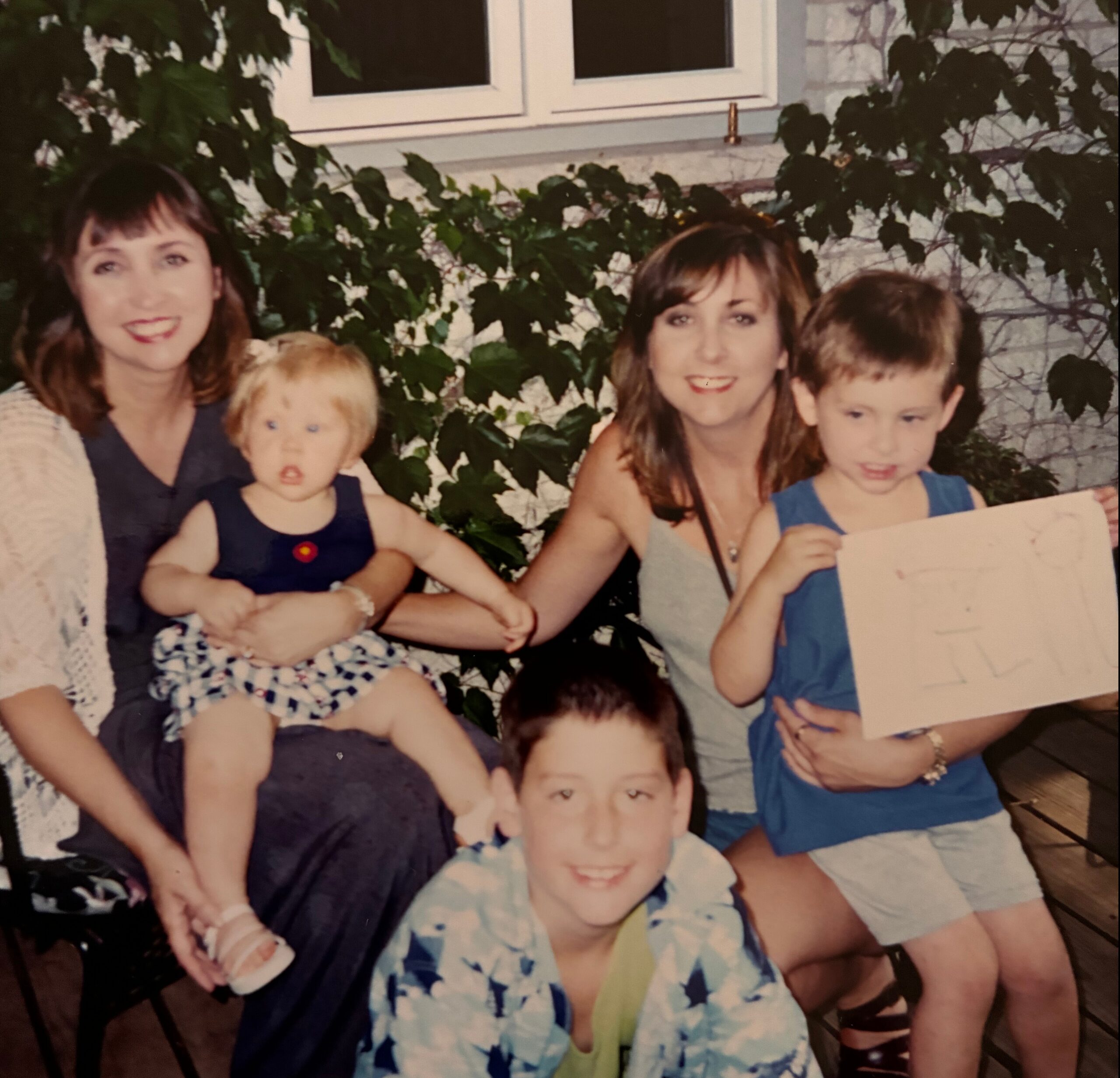 Family group photo outdoors with adults and children smiling in front of a vine-covered wall behind them.