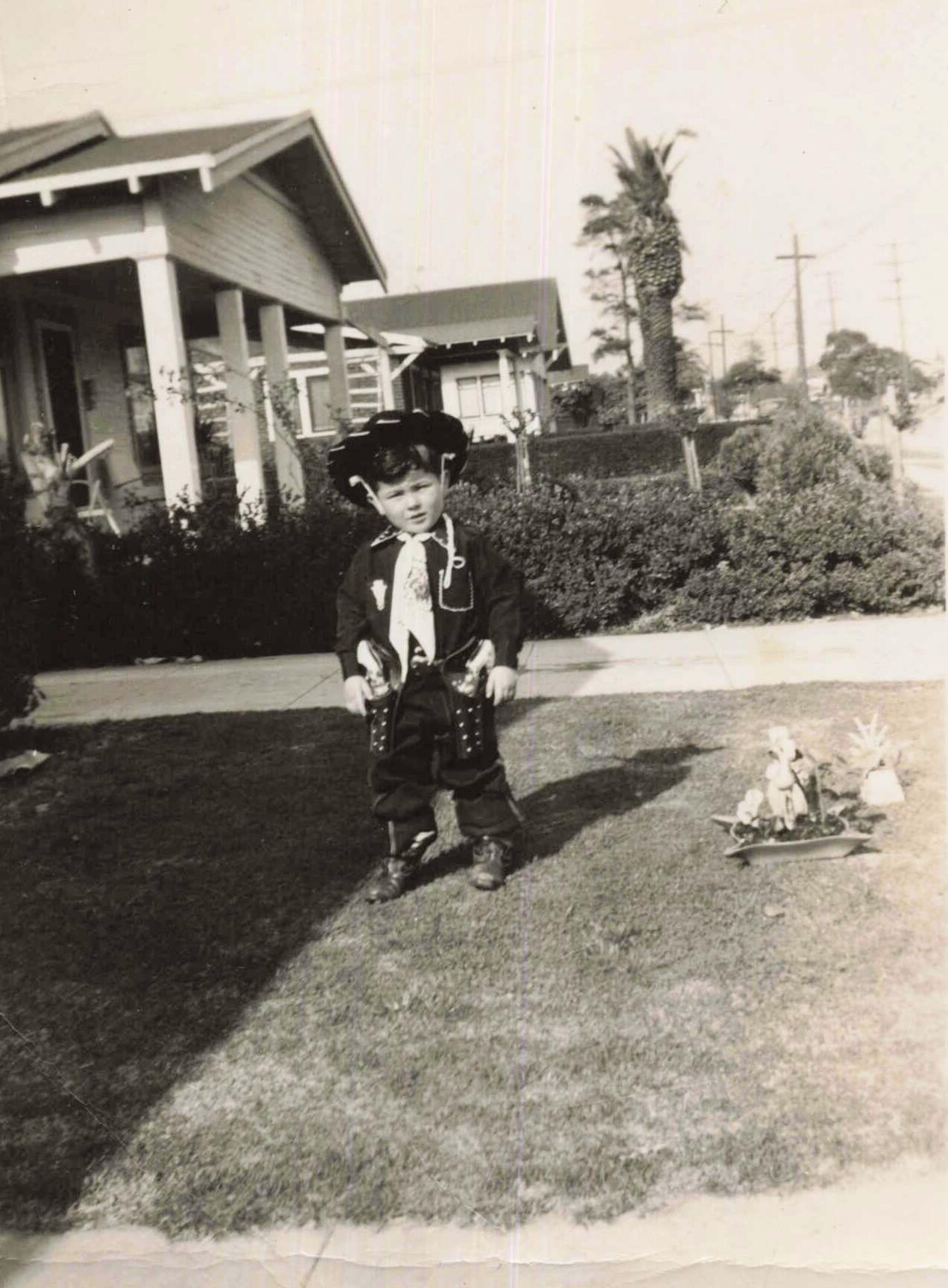 Young child in a cowboy outfit and hat standing on a lawn in front of suburban houses, with a small decorative plant in a tray to the right.