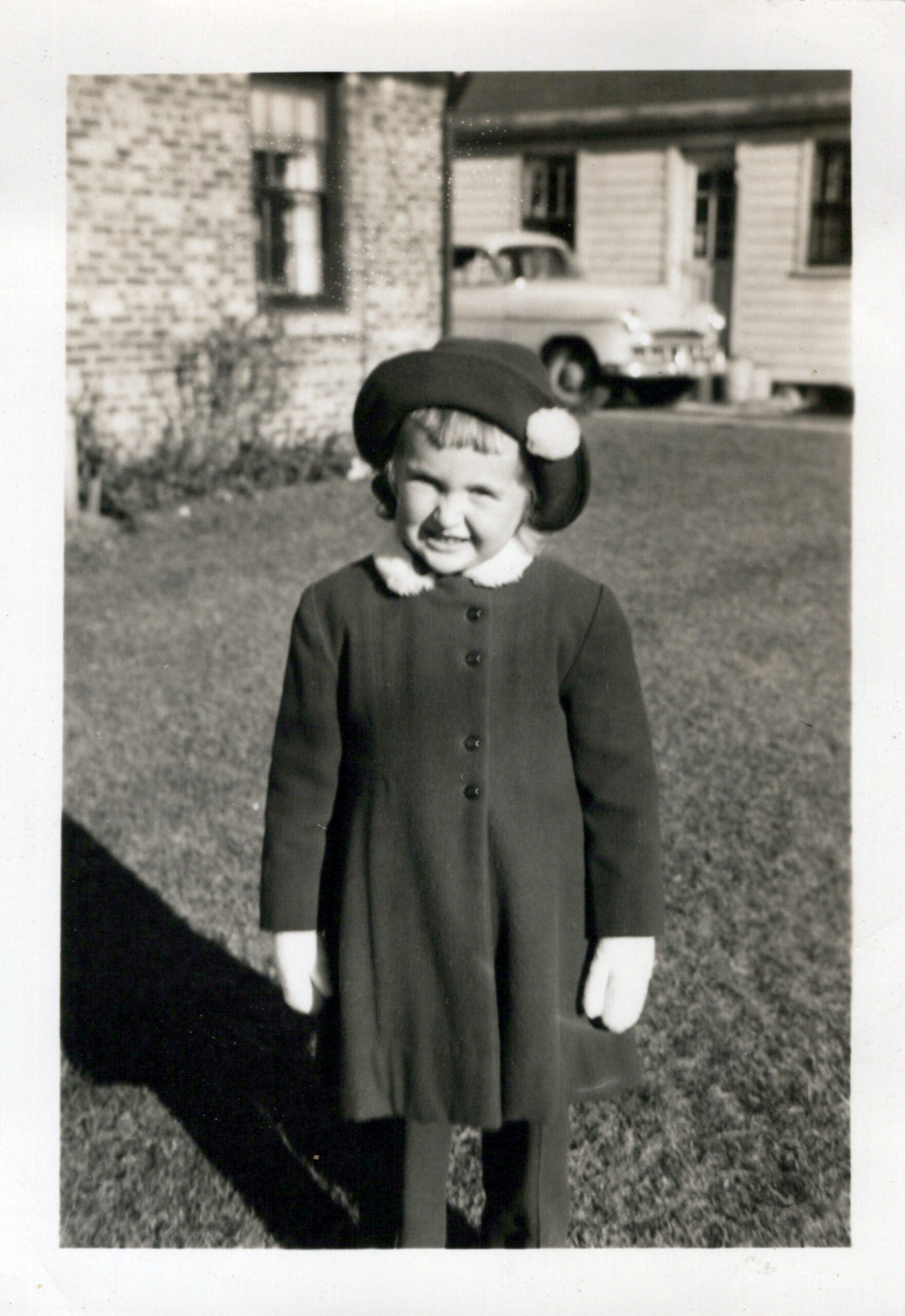 Smiling young girl in a dark coat and a hat with a pom-pom, standing on a lawn with a brick house and vintage car in the background inside a driveway setting.
