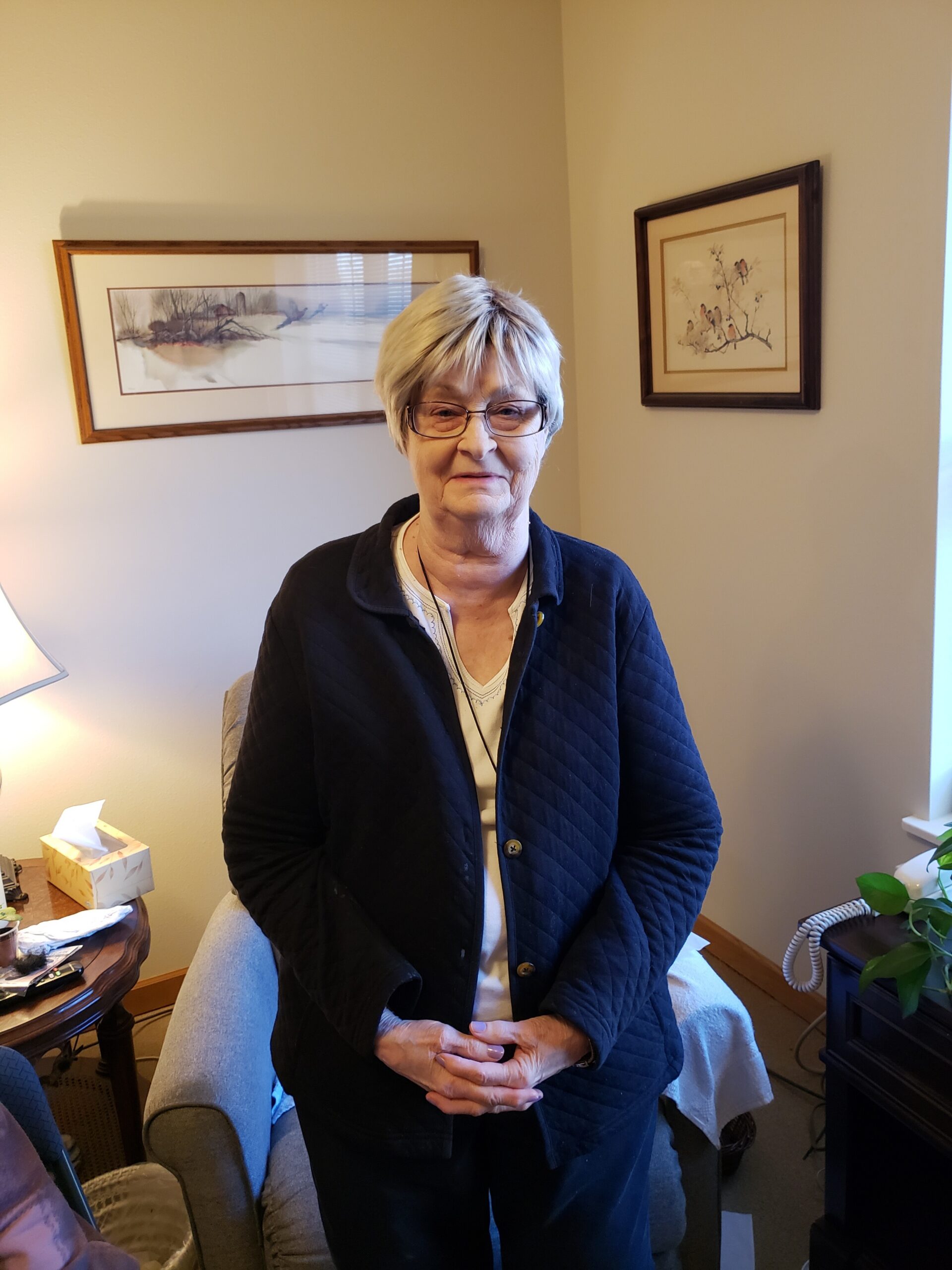 Older woman with short gray hair and glasses stands indoors in a beige-walled living room, wearing a dark quilted jacket.