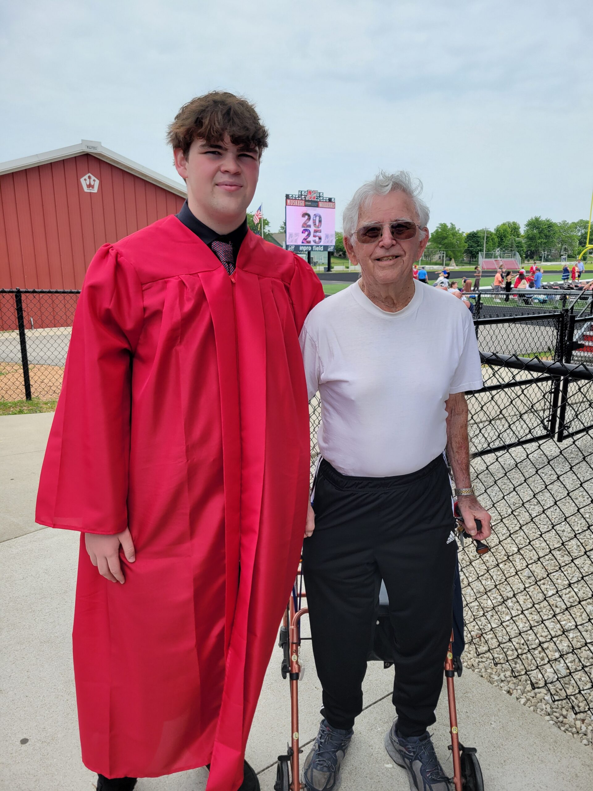 Young graduate in a bright red gown standing beside an elderly man in a white shirt at an outdoor sports field, both smiling.