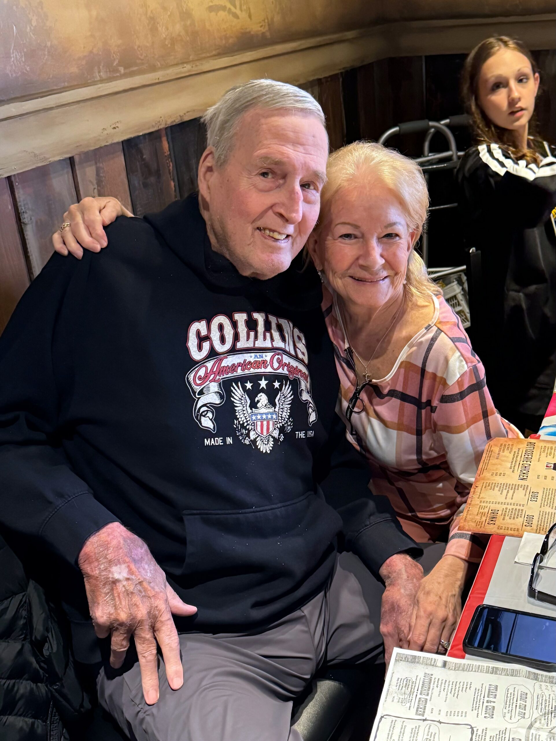 Smiling elderly couple posed together at a restaurant table, with menus visible on the table nearby.