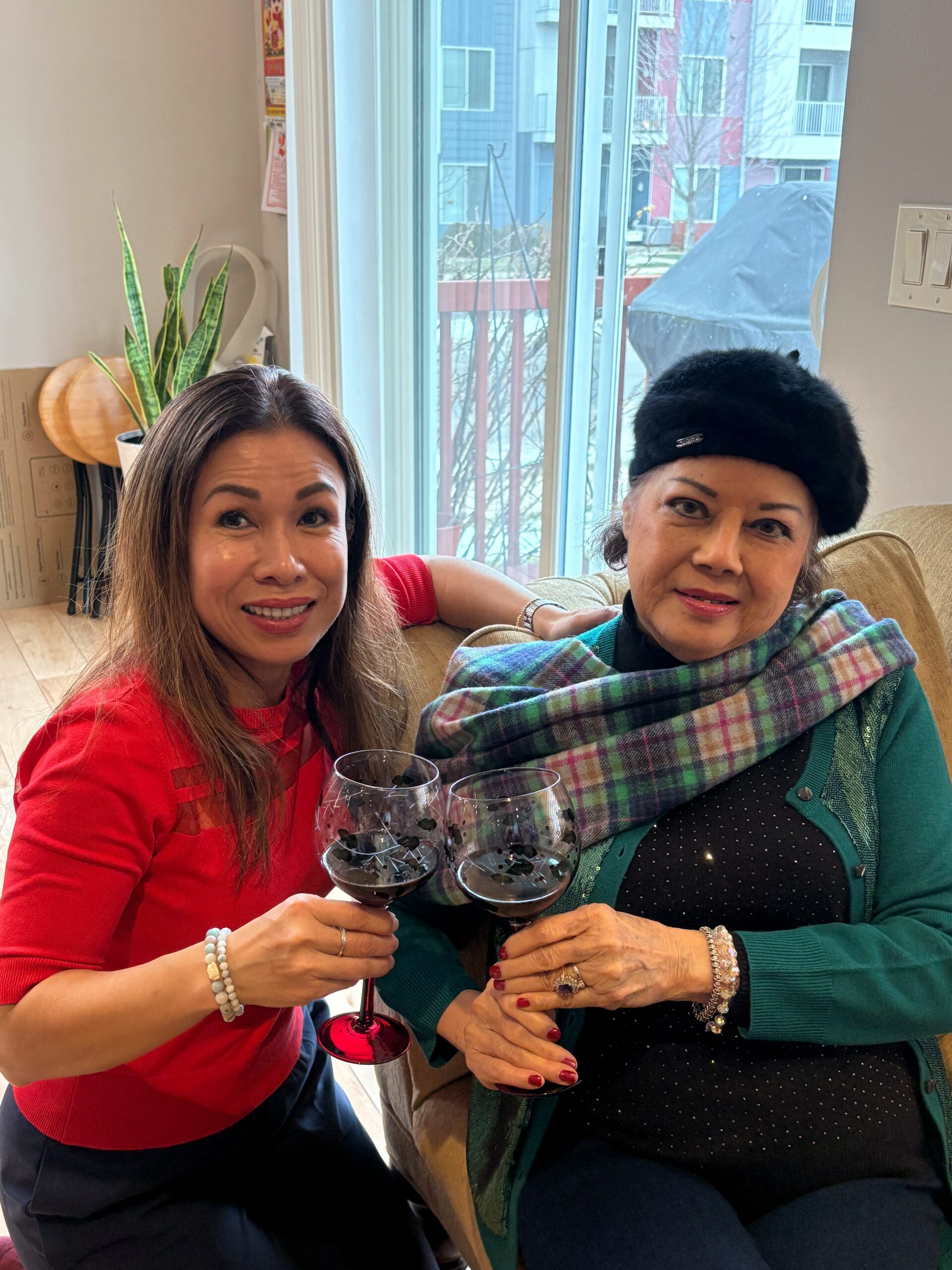 Two women toast with glasses of red wine in a living room; one wearing a red sweater, the other in a green cardigan and black hat.