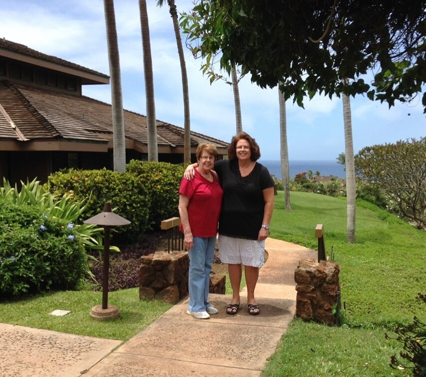 Two women standing arm-in-arm on a garden path with palm trees and a distant ocean view.