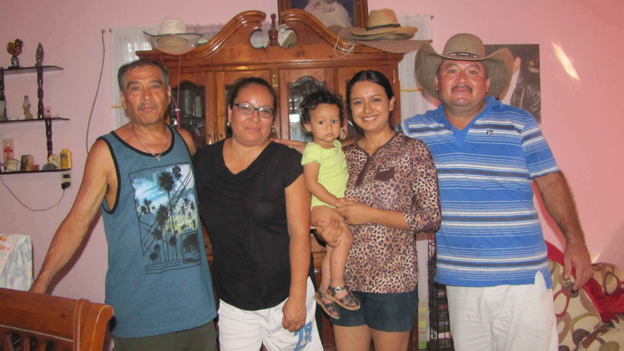 Five people posing indoors in a pink room: two men, two women, and a toddler held by a woman in a leopard print top, with a wooden cabinet behind them.