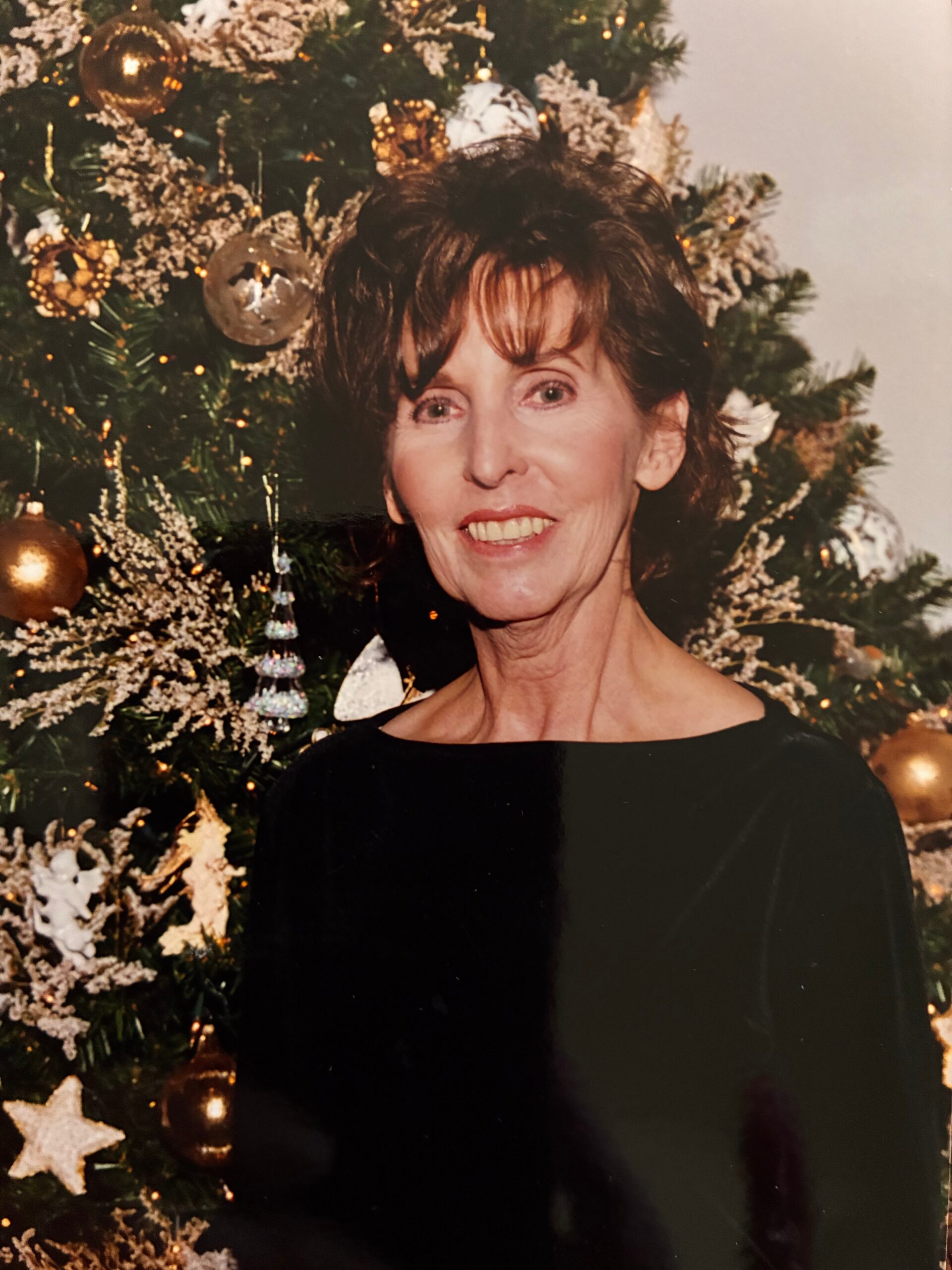Smiling woman with short brown hair in a black top posing in front of a Christmas tree adorned with gold ornaments and snowy accents.