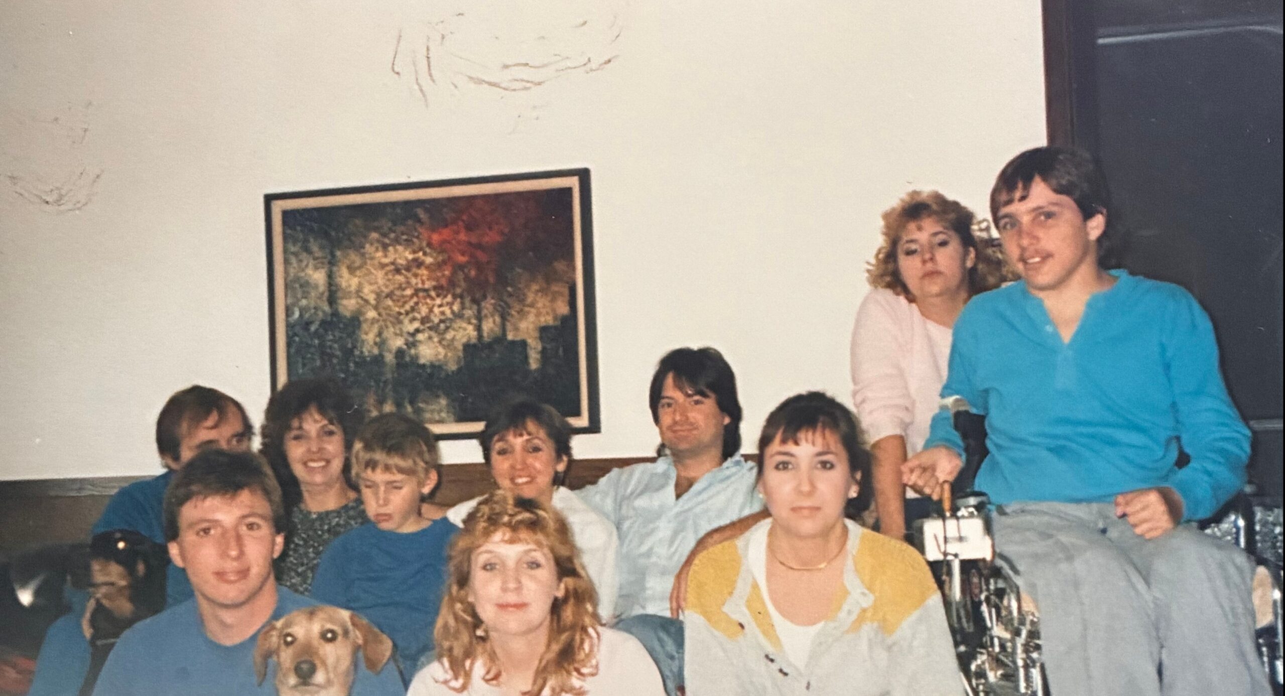 A group of people posing for a casual photo in a living room, with a dog in the foreground and a framed painting on the wall behind them.