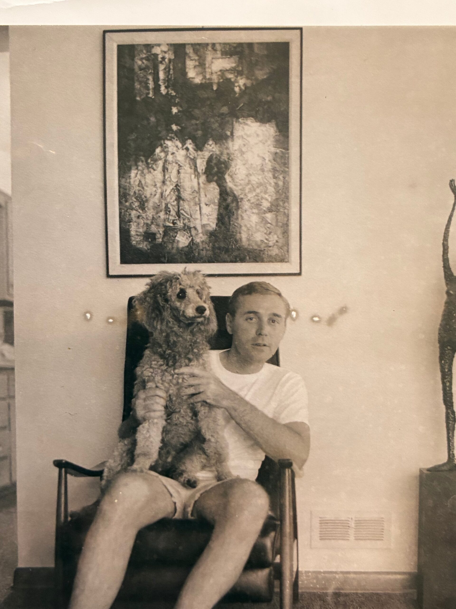 Man seated in a chair, holding a large fluffy dog on his lap, in a room with a framed abstract painting on the wall behind him.