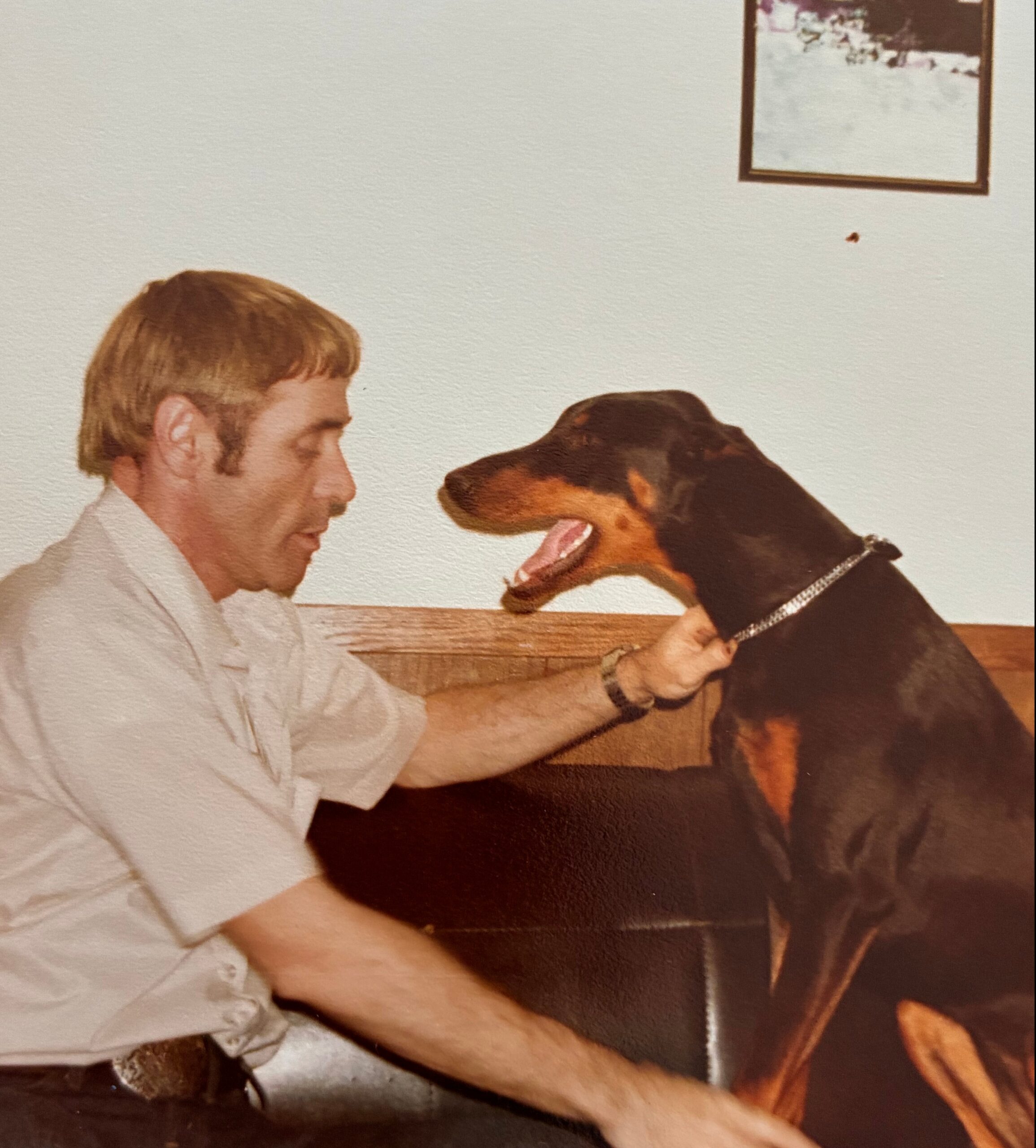 Man in a light shirt shares a close moment with a large brown and black dog, noses touching indoors on a wooden surface in the background.