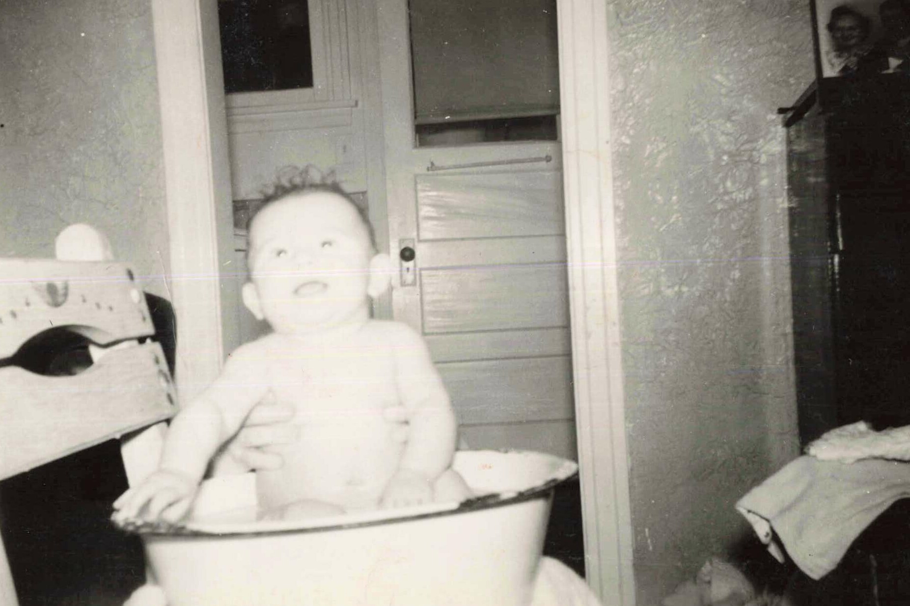 Black-and-white photo of a baby sitting in a metal tub indoors, with a doorway and a partially visible figure in the background.