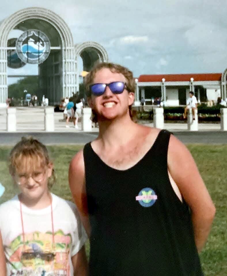 Smiling man in a black sleeveless shirt wearing blue sunglasses beside a girl in a white shirt on a grassy area, with a futuristic arched building in the background.
