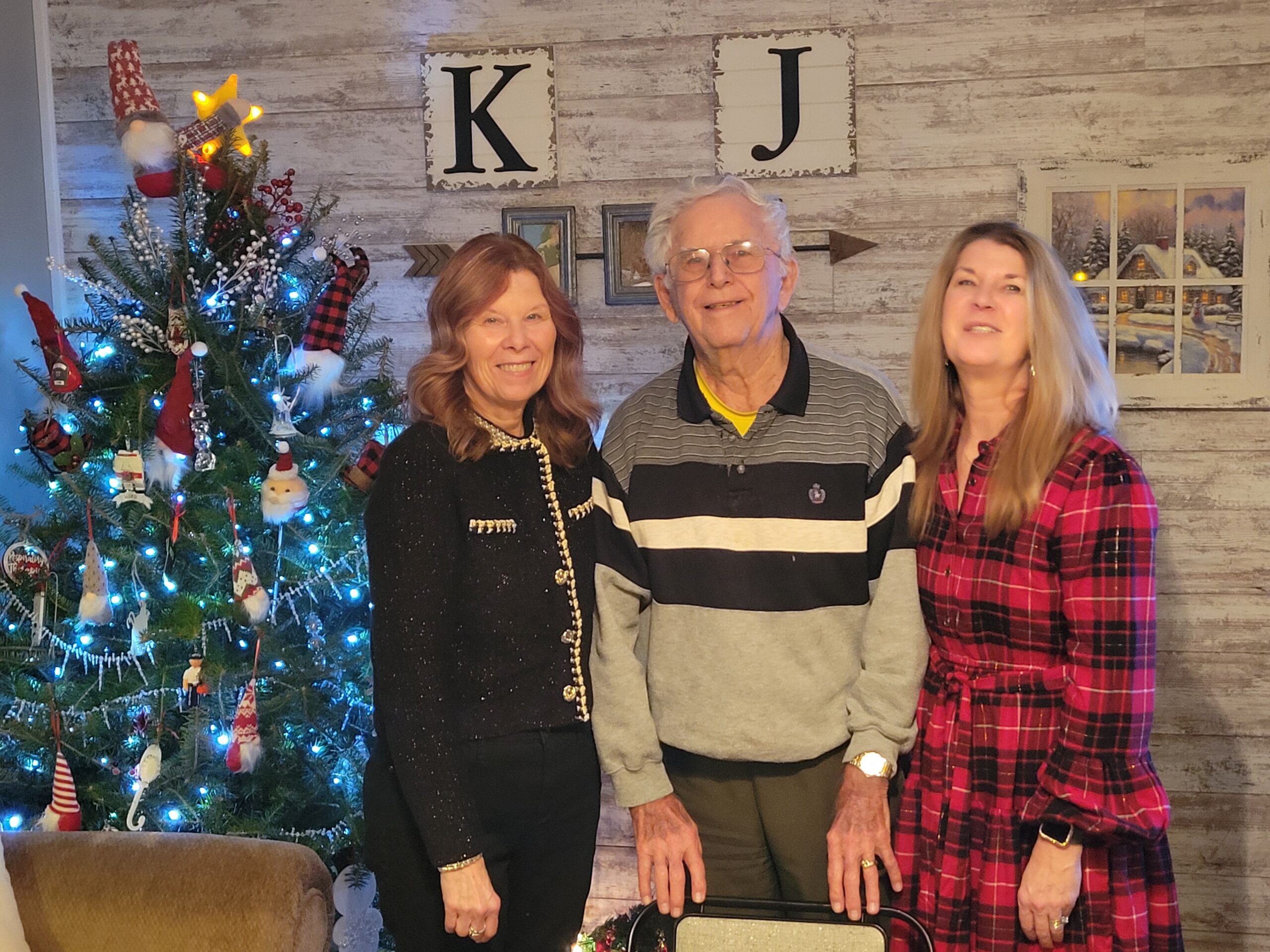 Three adults pose together indoors beside a decorated Christmas tree and a rustic wall with K and J blocks.