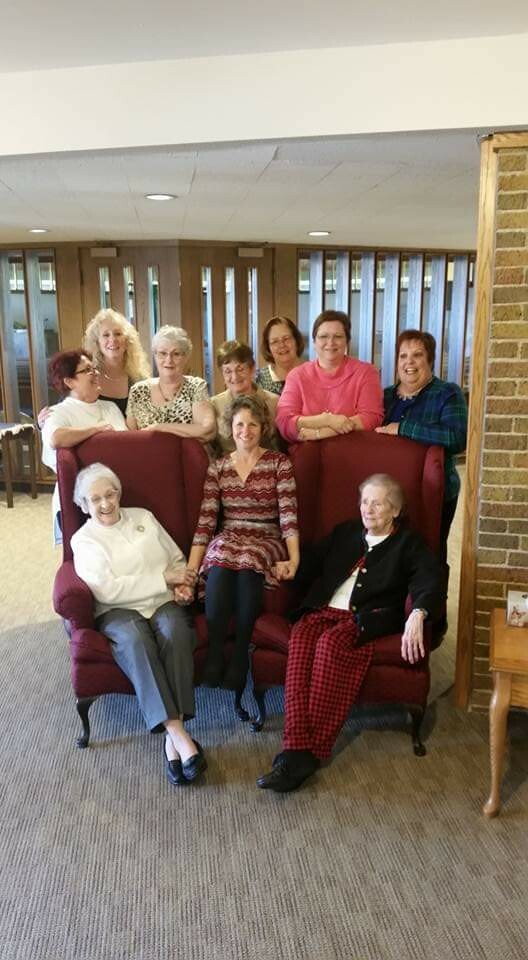 Group of nine women posing together indoors, two elderly women seated on a red sofa in front while others stand behind them in a common room.