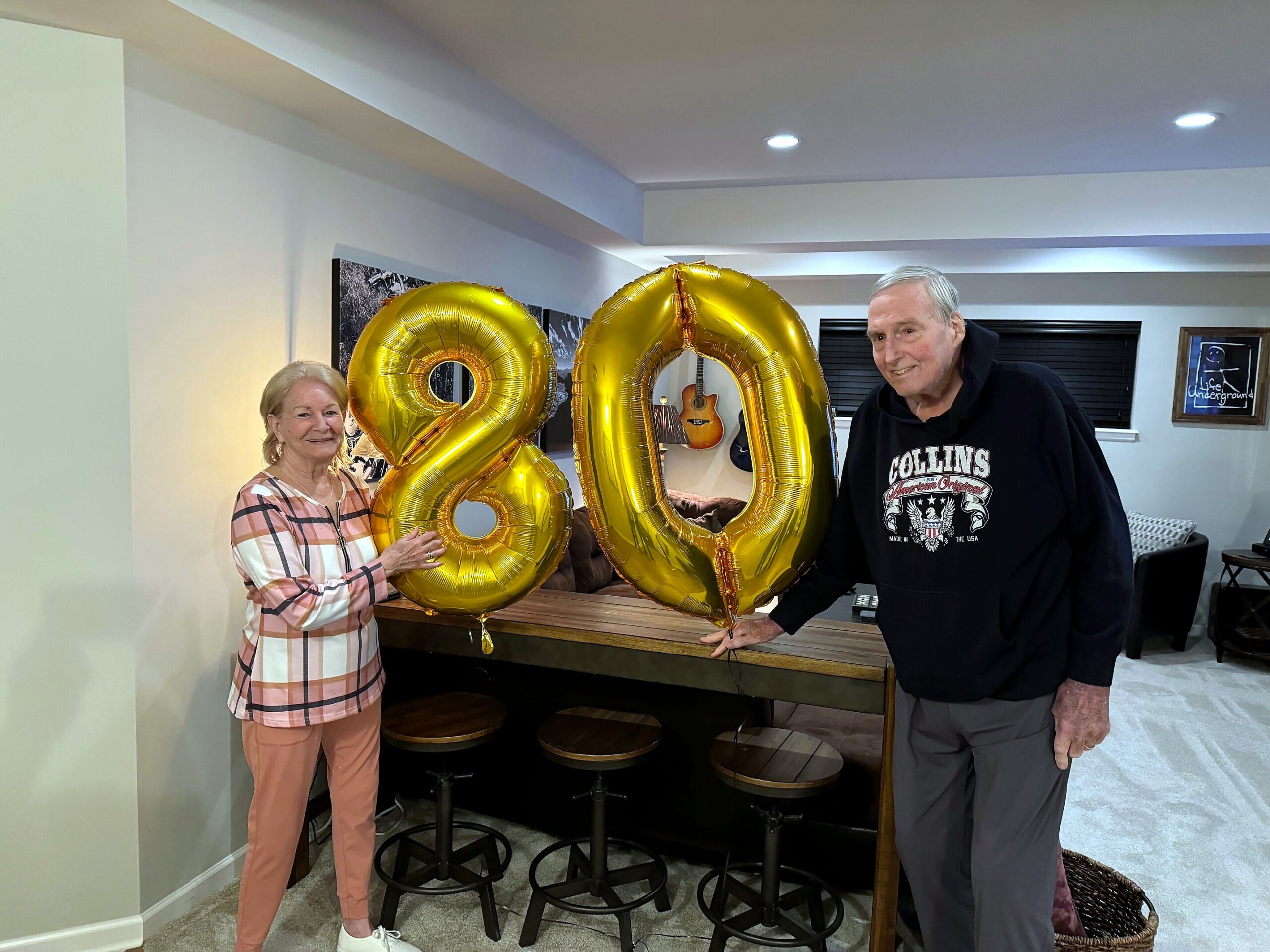 Two elderly adults indoors, smiling, holding large gold '80' balloons for a birthday celebration.