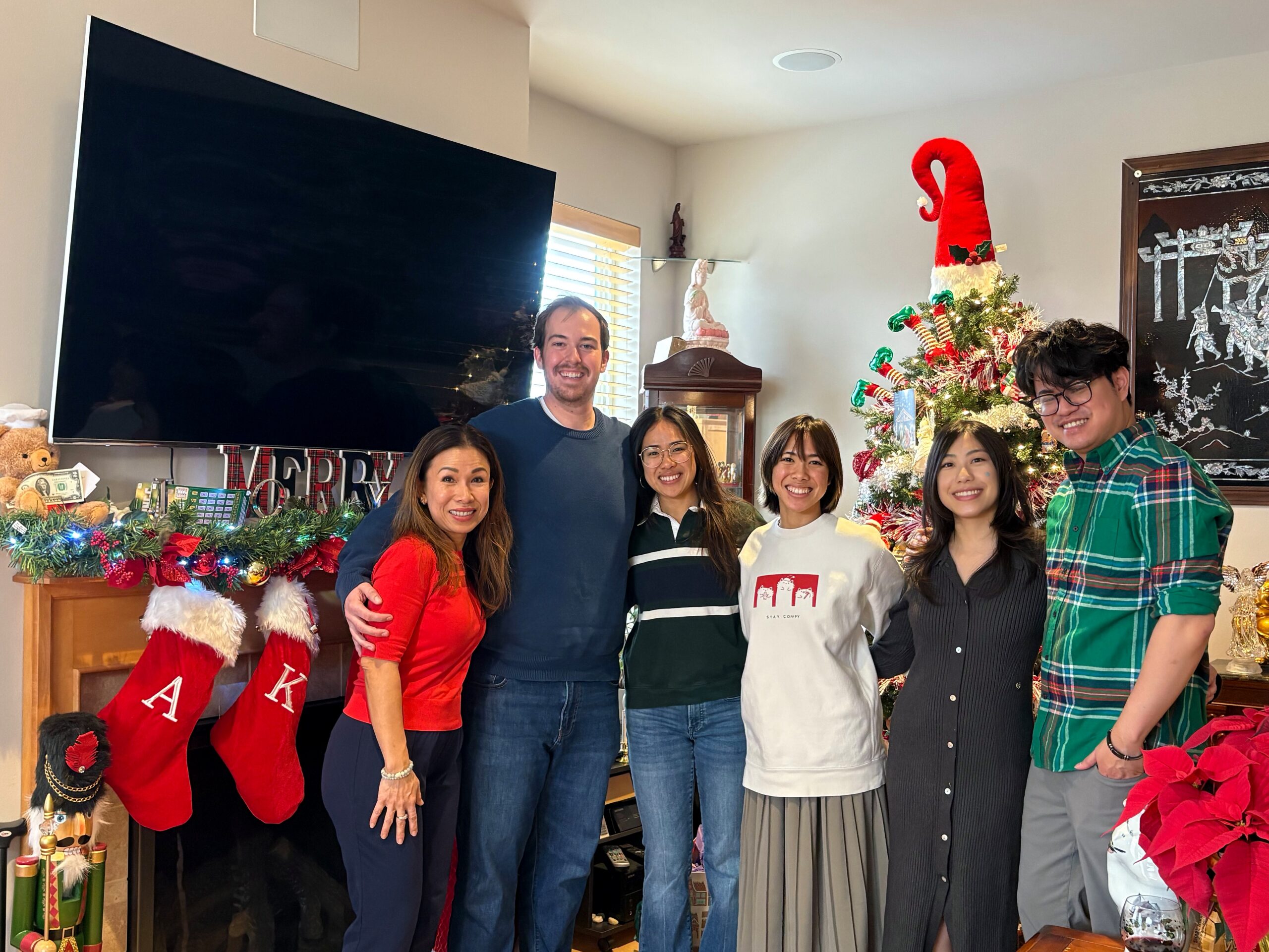 Group of adults posing for a holiday photo indoors with a decorated Christmas tree and stockings in the background.