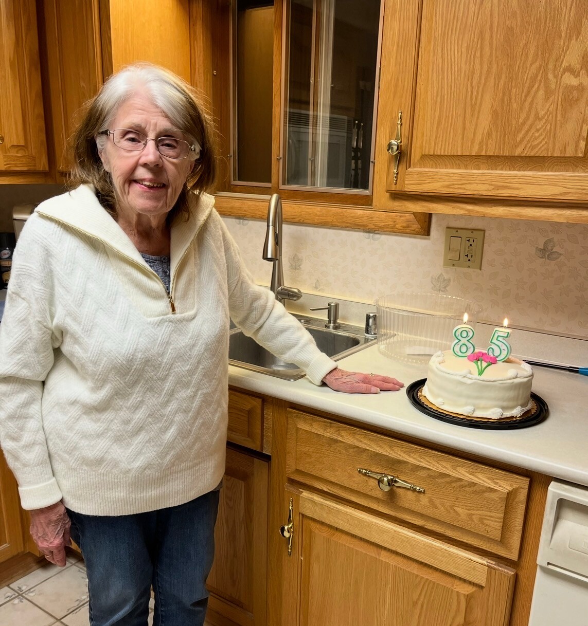Elderly woman in a white sweater stands by the kitchen counter beside a birthday cake with '85' candles.