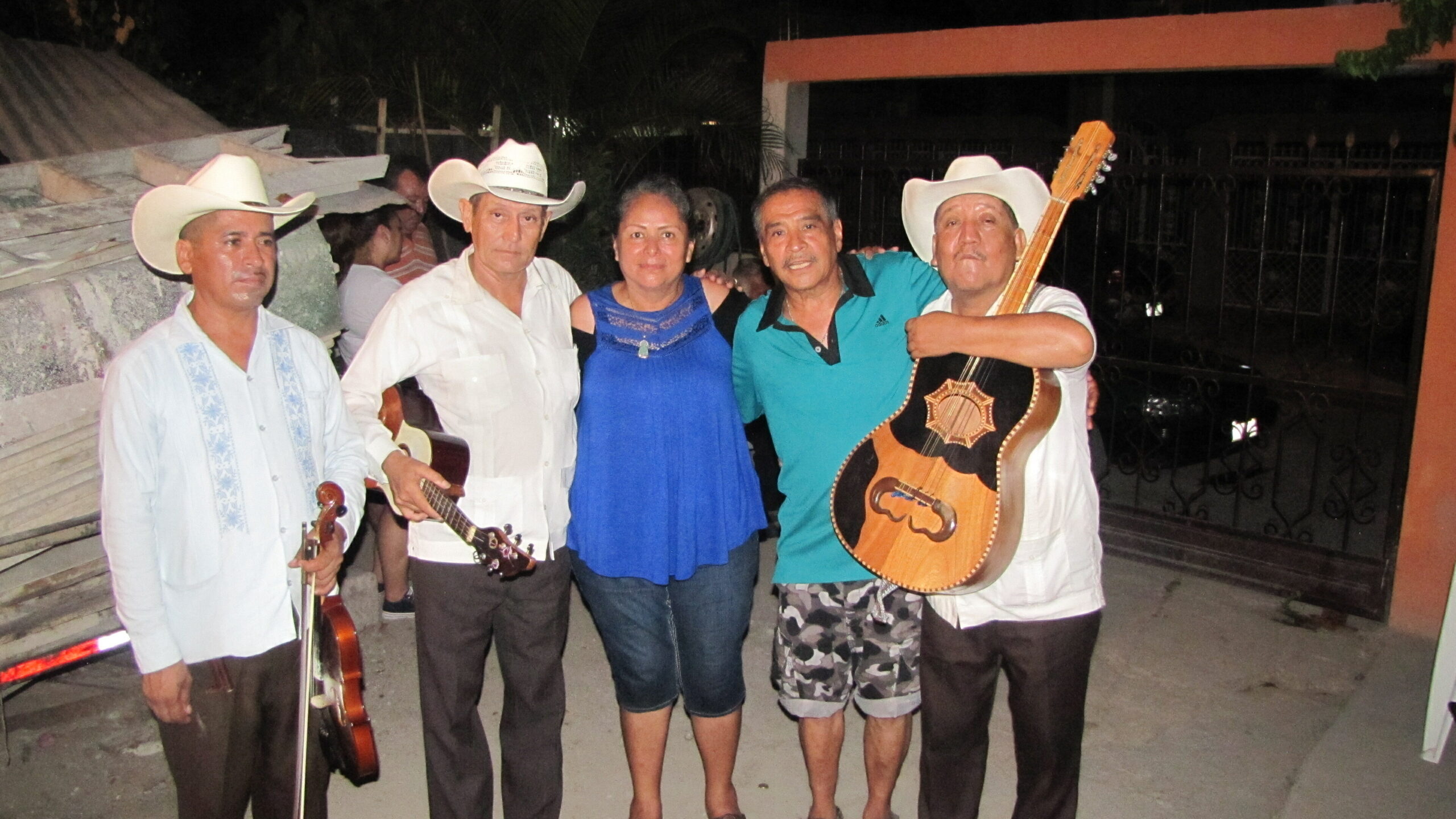 Five adults posing outdoors at night; two men wear cowboy hats and hold guitars, a woman in blue stands between them.