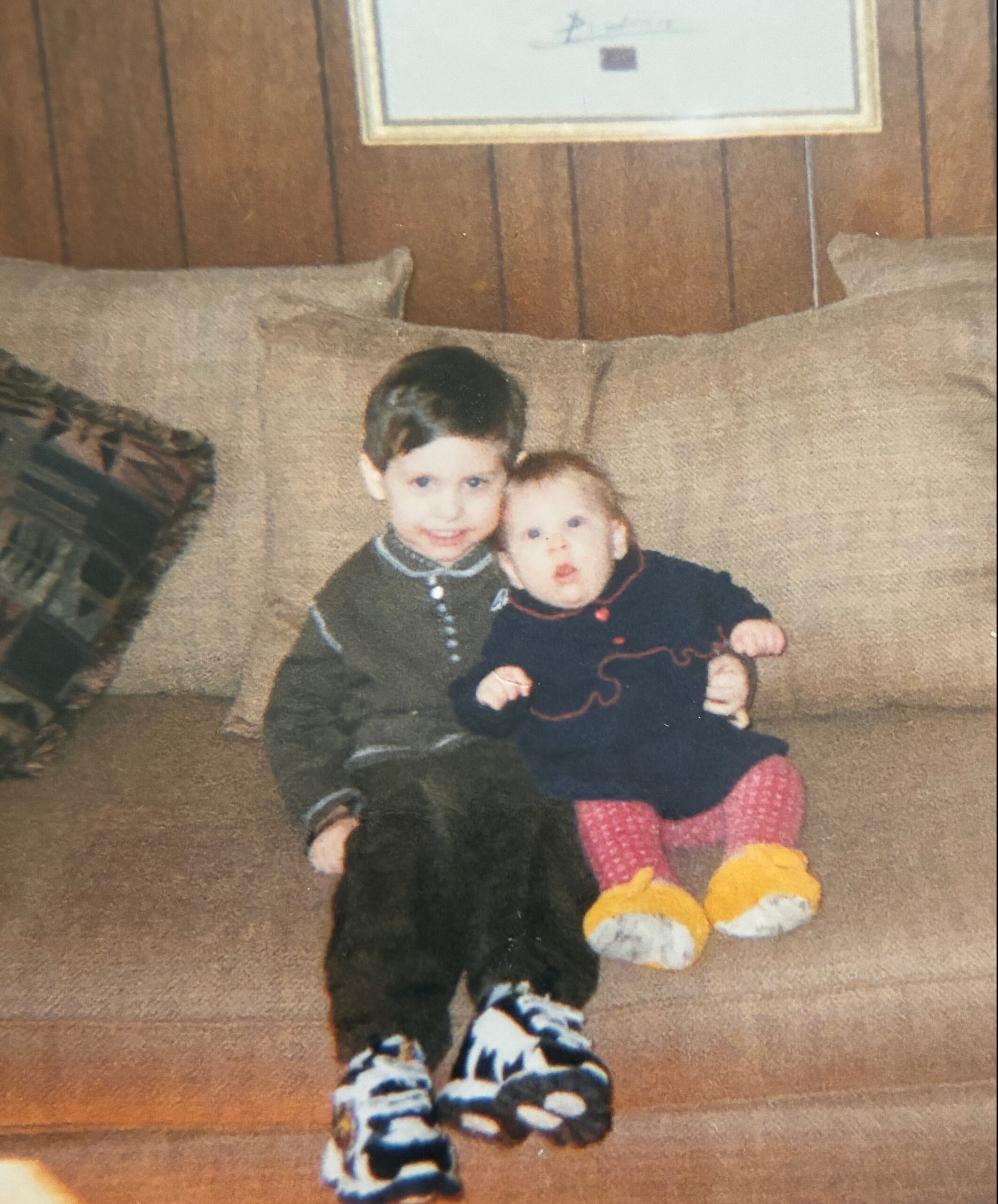 Two young children sit close together on a brown sofa indoors, with yellow plush toys at their feet and a wood-paneled wall behind them.