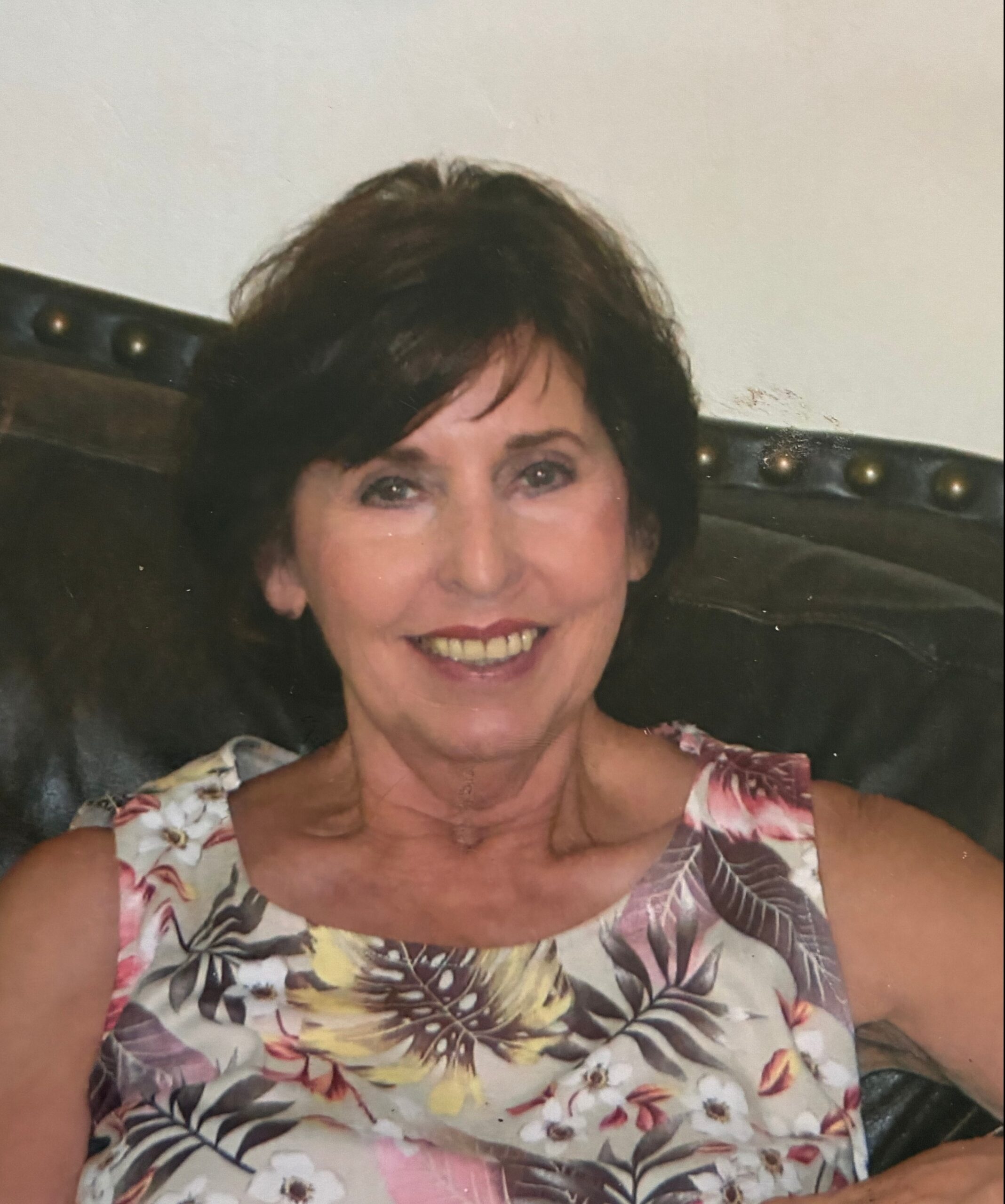 Smiling woman with short dark hair wearing a colorful floral dress, seated against a dark upholstered headboard.