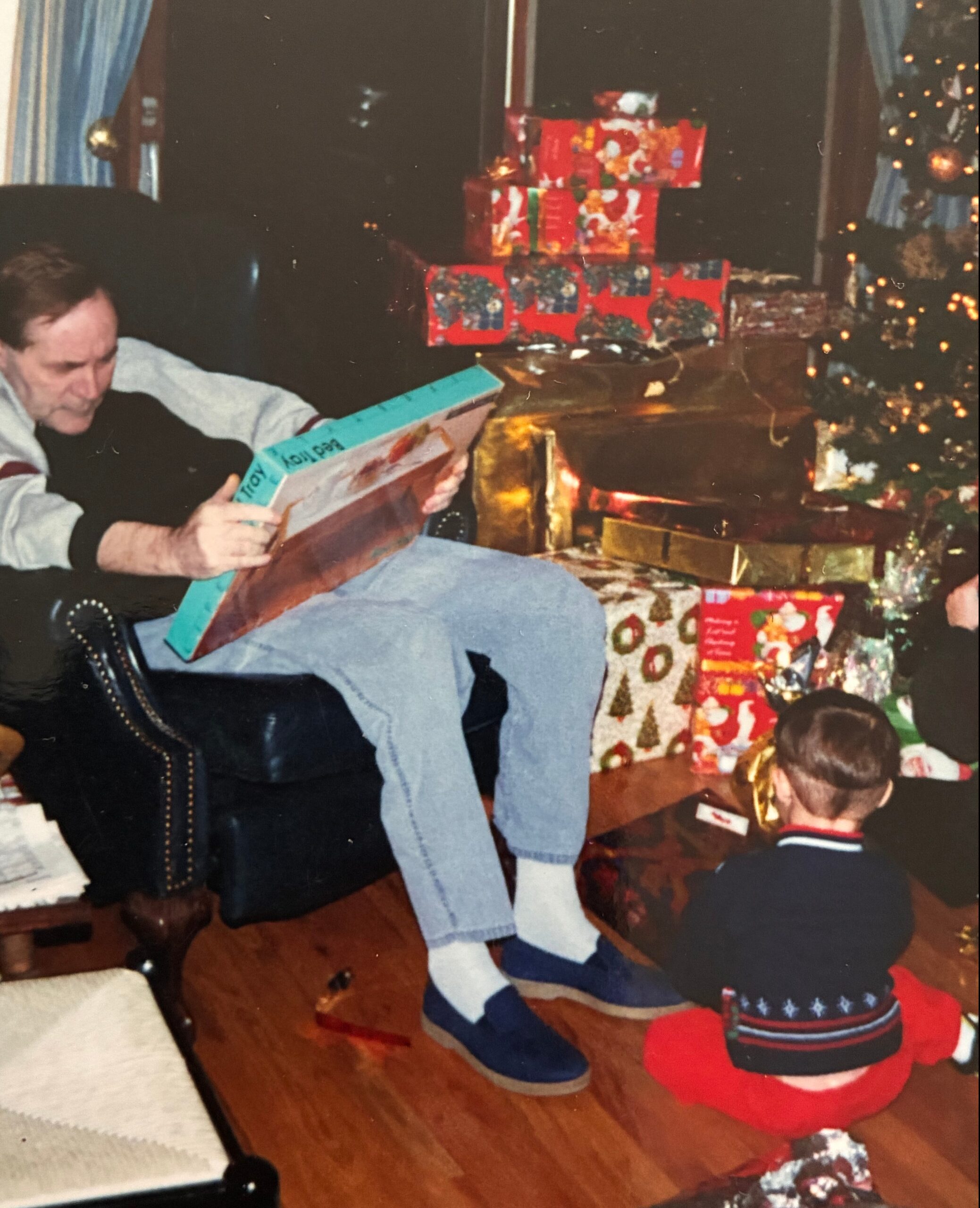 Man in a chair unwrapping a large blue gift while a child sits nearby under a Christmas tree with wrapped presents.