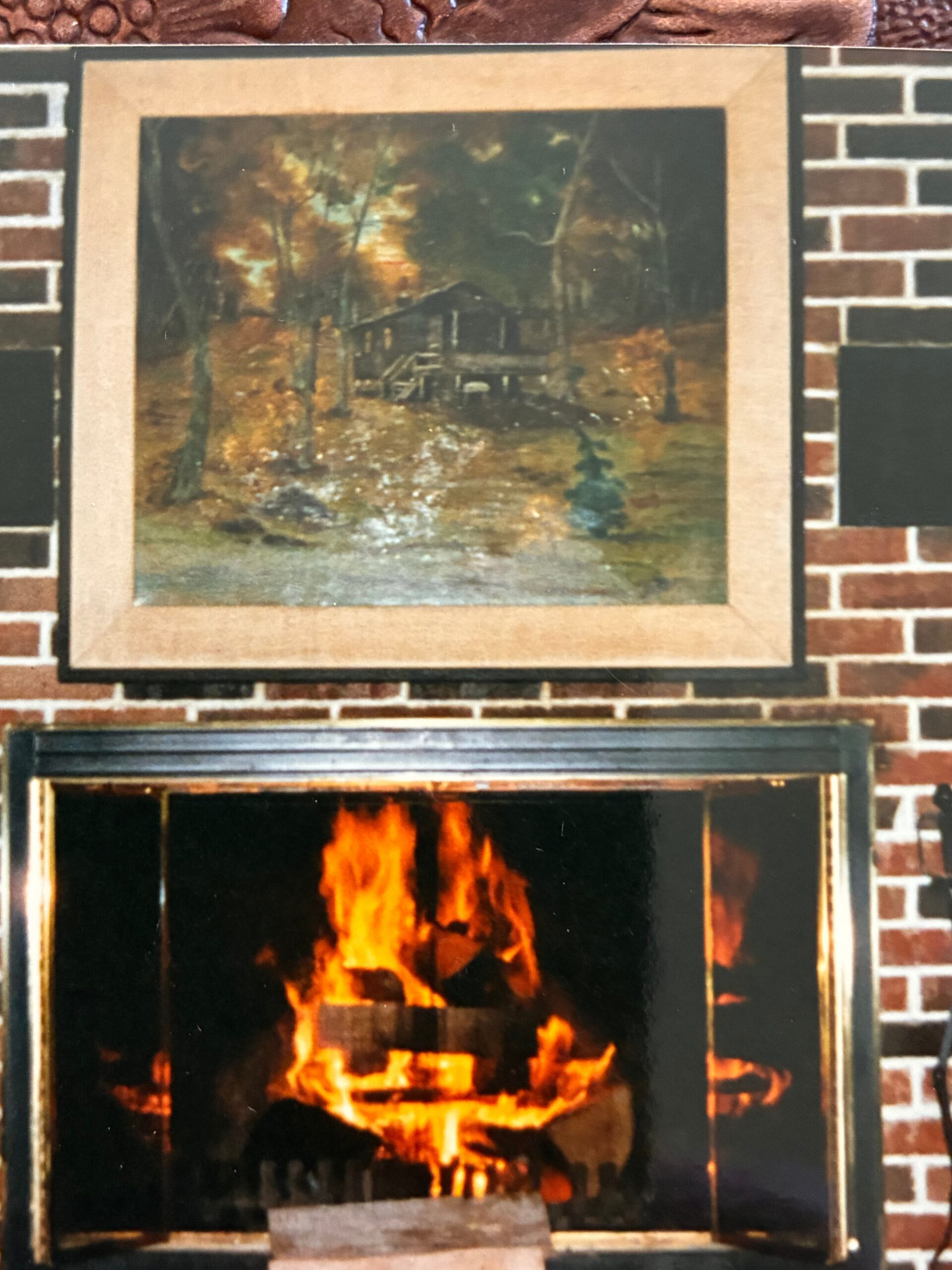 Framed landscape painting above a brick fireplace mantle, in a dimly lit living room setting