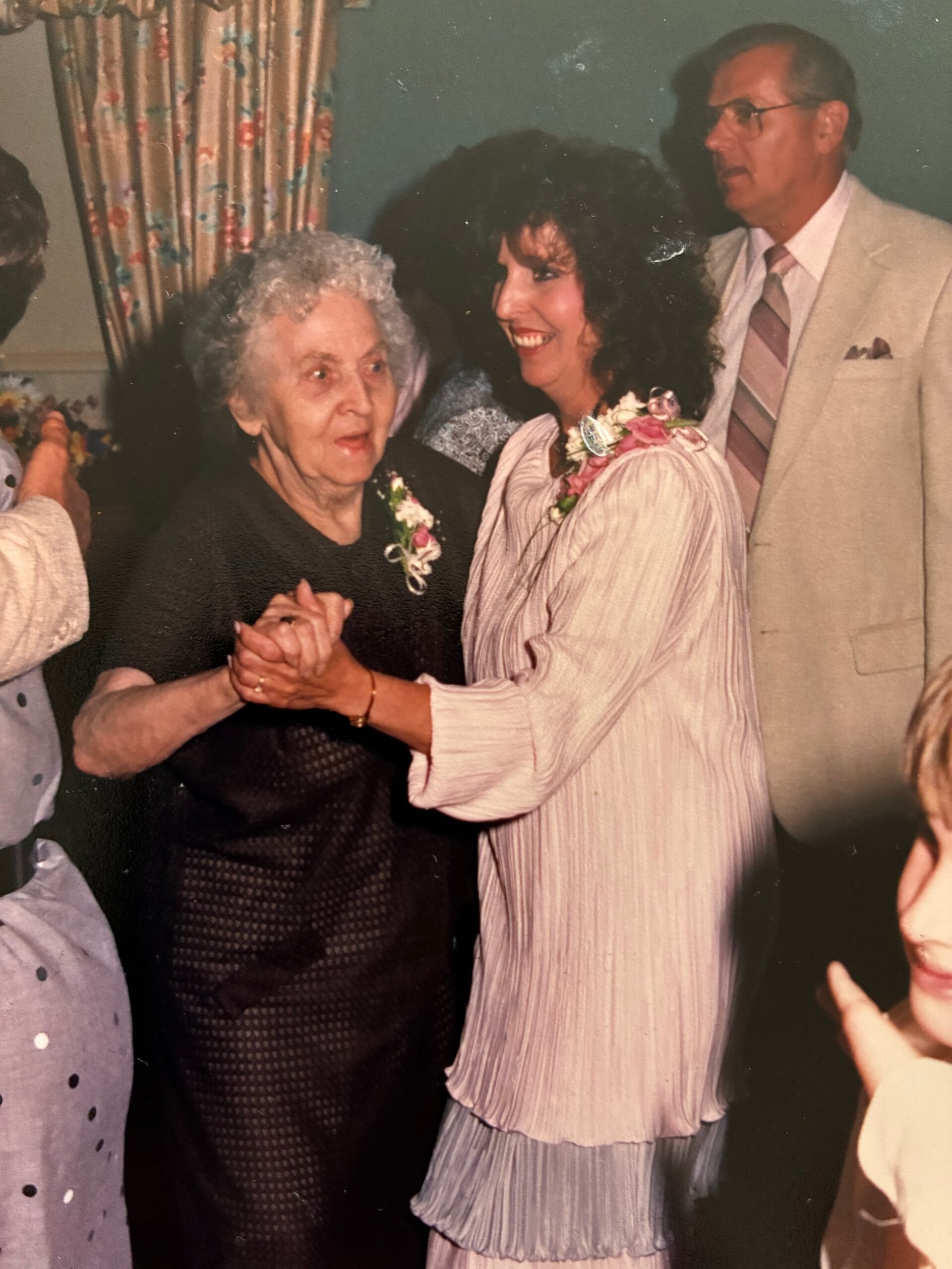 Two women dance together, smiling and holding hands at a celebration, with a man in a beige suit in the background.