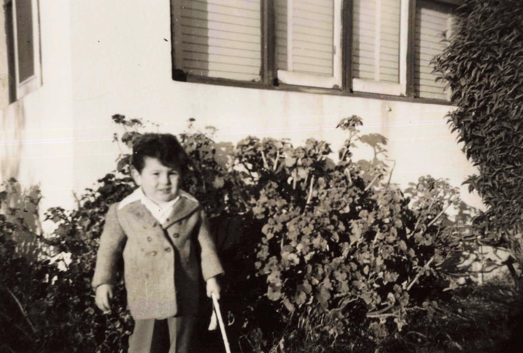 Young girl in a coat standing in a garden with a hedge, in front of a house wall and windows behind her.