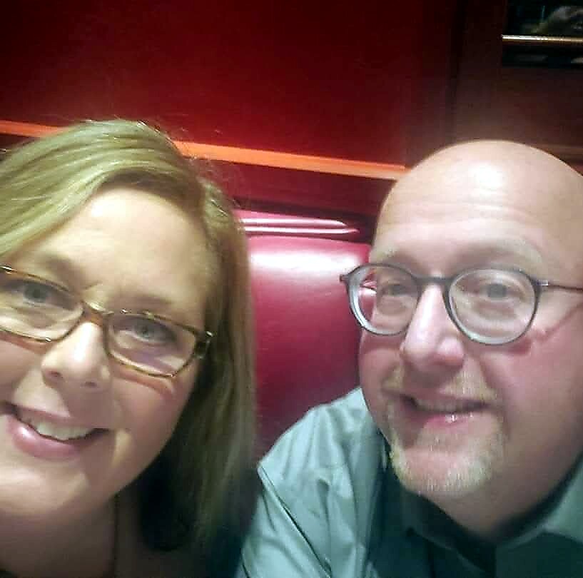 Two smiling adults sit close together in a red booth at a restaurant, looking at the camera (woman on left with glasses, man on right with glasses).