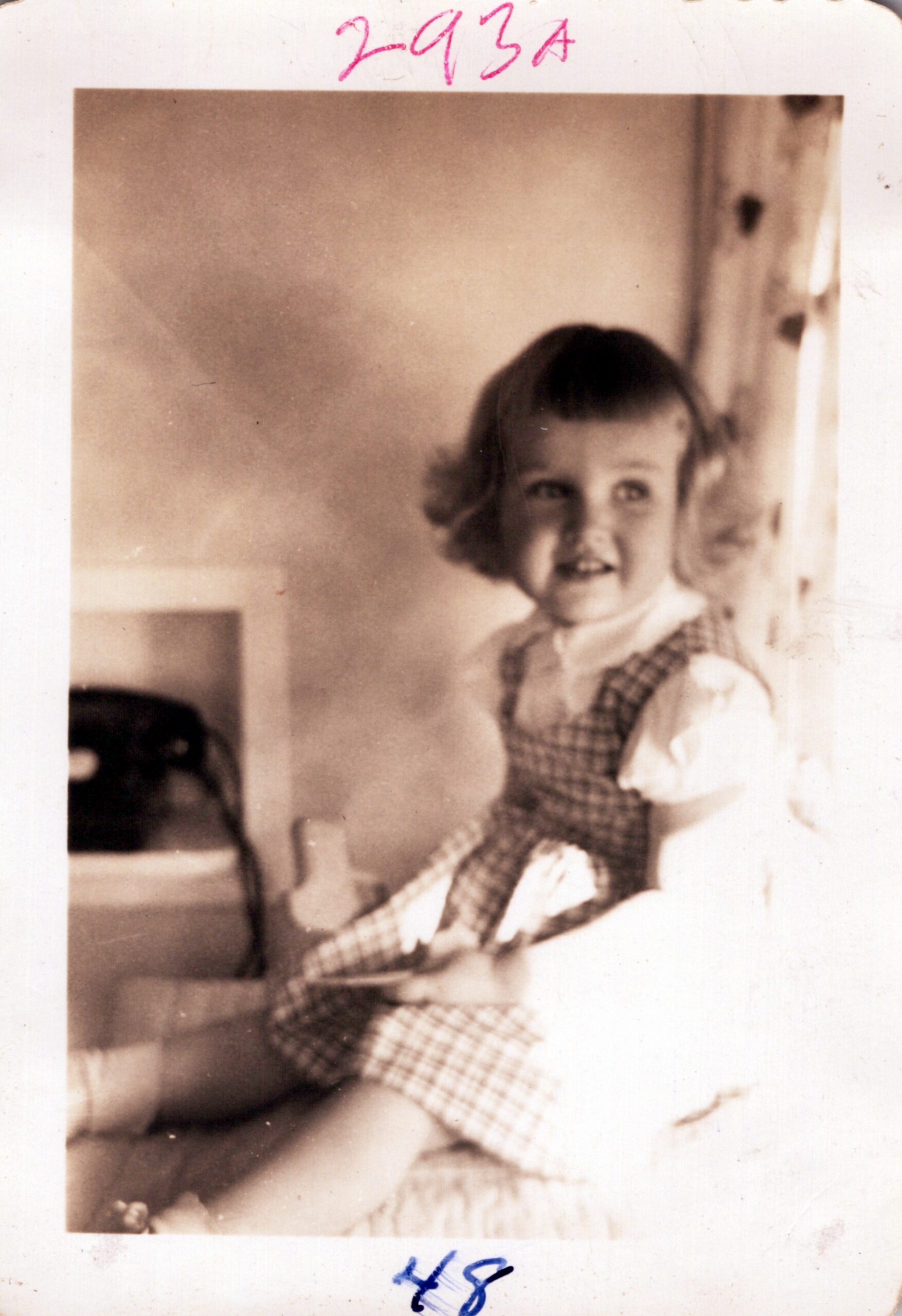 Smiling young girl with short dark hair wearing a plaid dress, indoors, looking toward the camera.