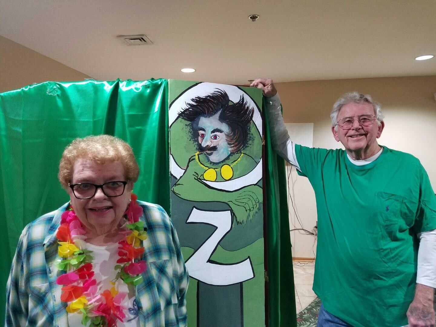 Two seniors posing beside a cartoon wizard cutout in a photo booth with a green backdrop behind them, both smiling.