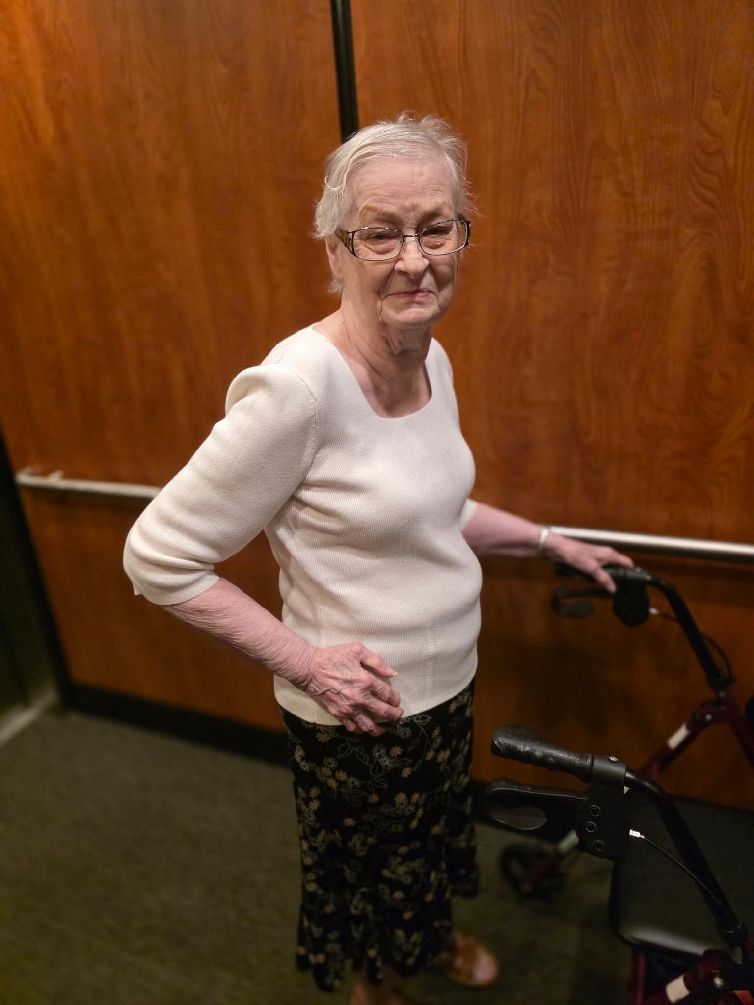 Older woman with short white hair and glasses stands beside a walker in a wood-paneled hallway, wearing a light sweater and patterned skirt, with a gentle smile.