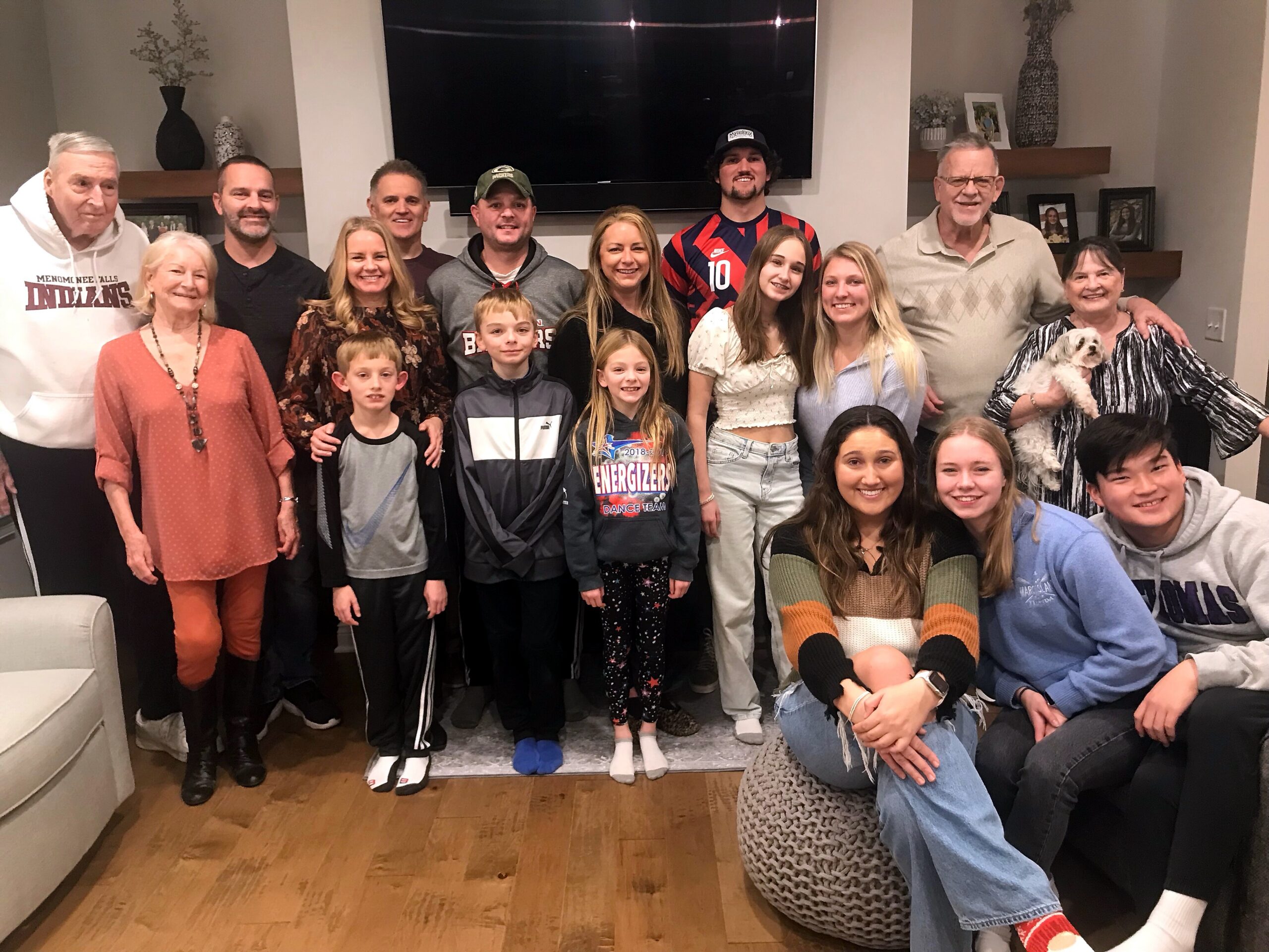 Group of adults and children posing together in a living room, with a large TV on the wall behind them and a dog appearing in one arm of a woman on the right.