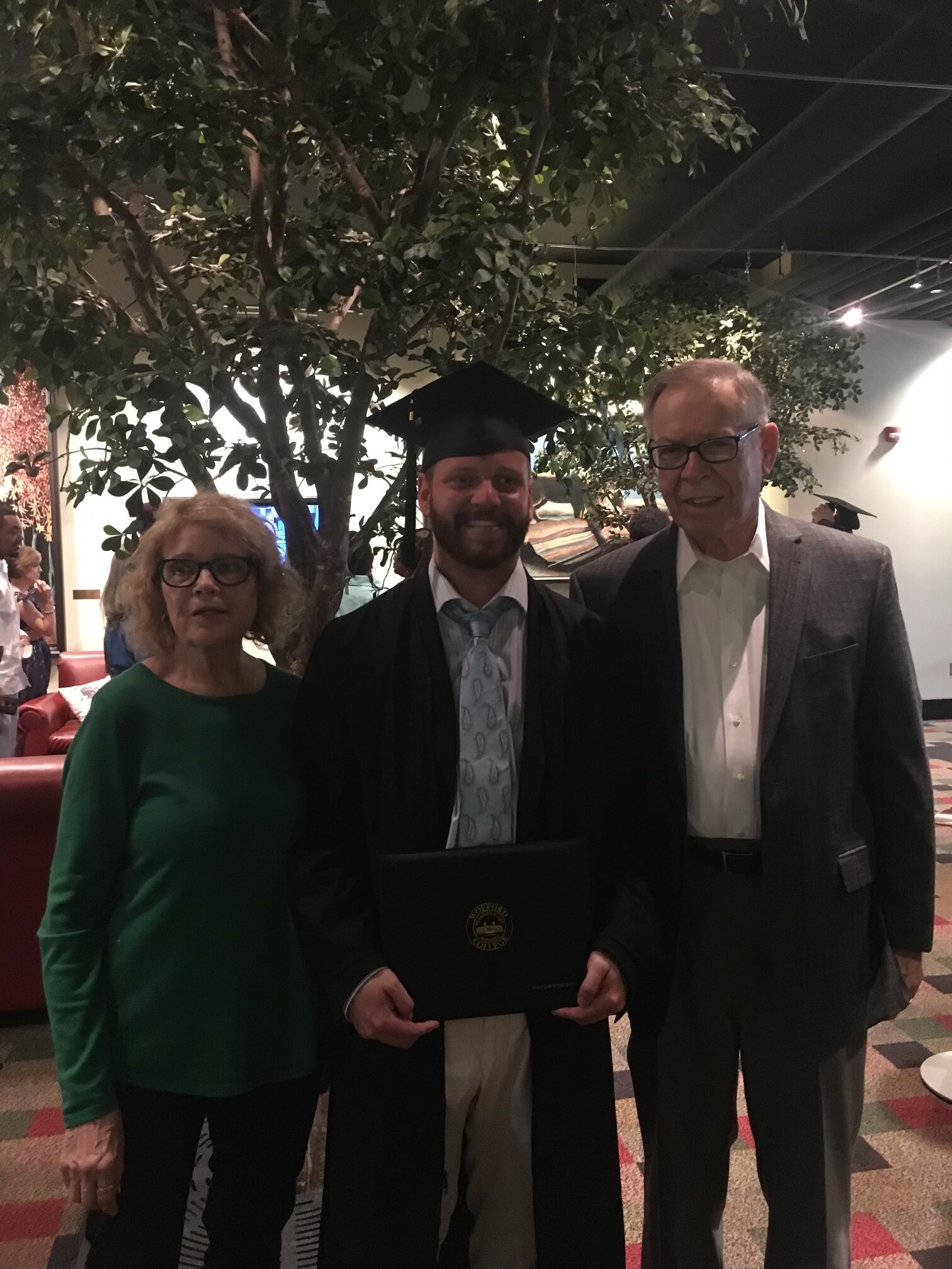 Graduate in cap and gown posing with two adults indoors, greenery and soft lighting in the background.