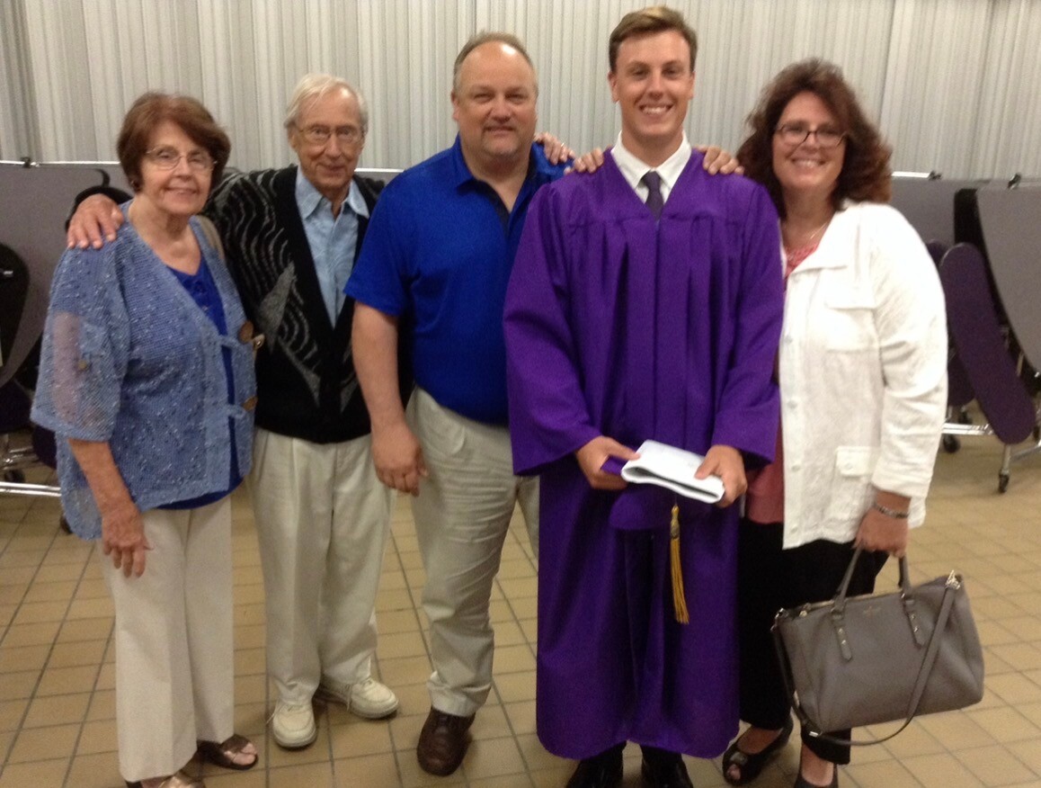 Graduate in a purple gown holding a booklet, posing with family indoors.