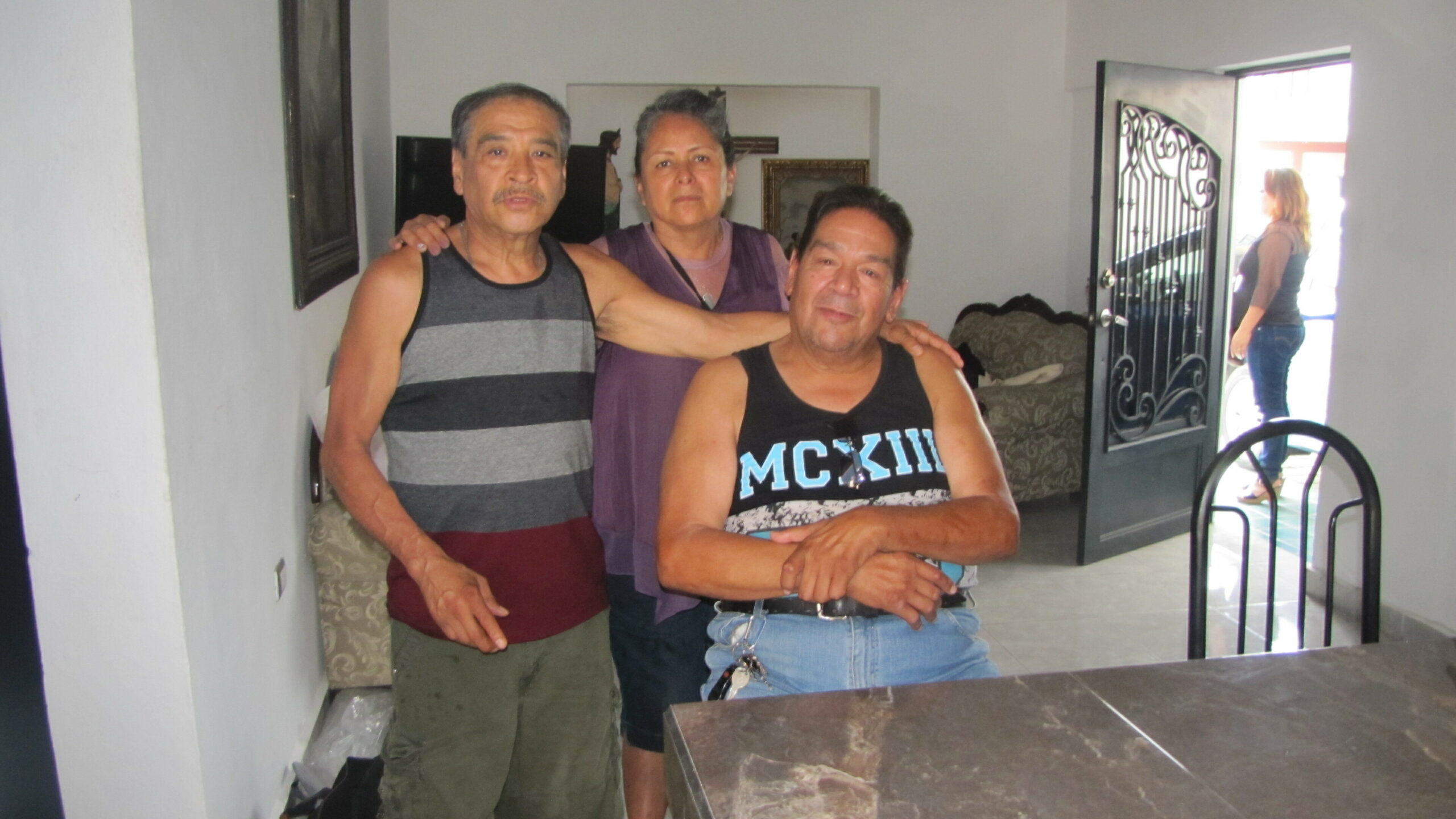 Three adults pose together in a living room, two men flanking a man in front with his arms crossed, a woman stands behind them in purple.