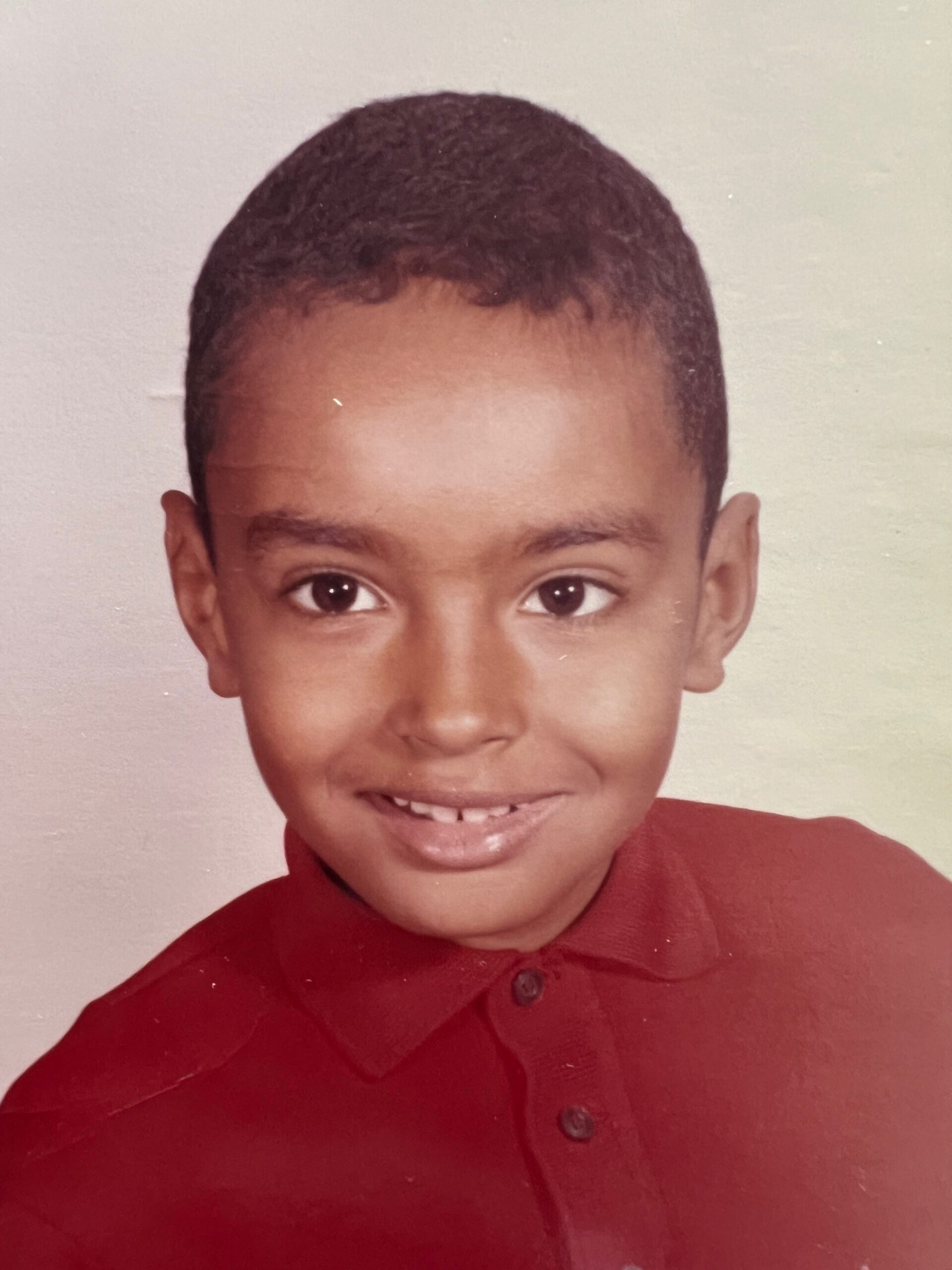 Smiling young boy with short dark hair wearing a red polo shirt, looking at the camera against a neutral background.