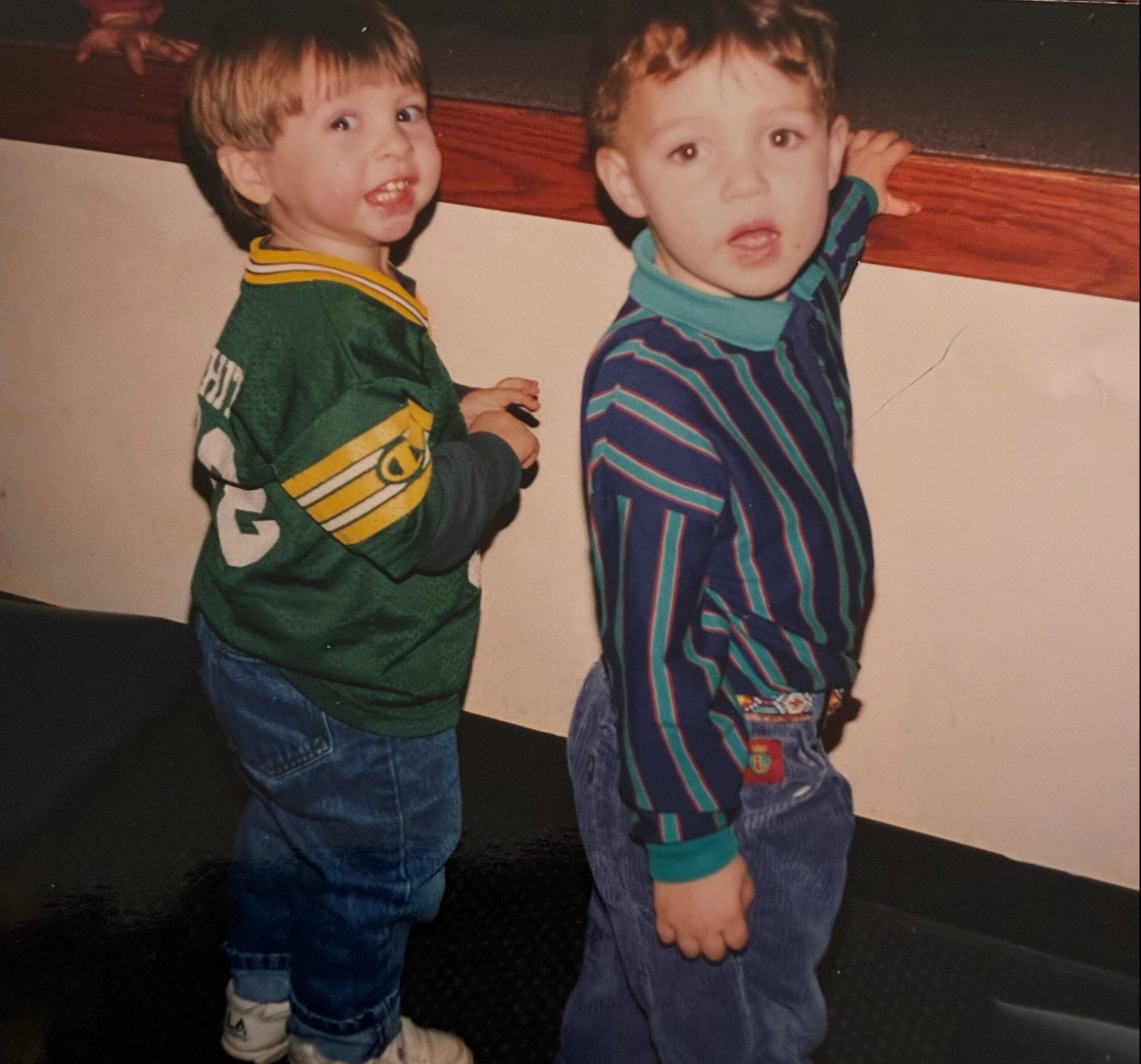 Two young boys stand side by side indoors, one in a green jersey and the other in a striped blue outfit, both looking at the camera with surprised expressions.