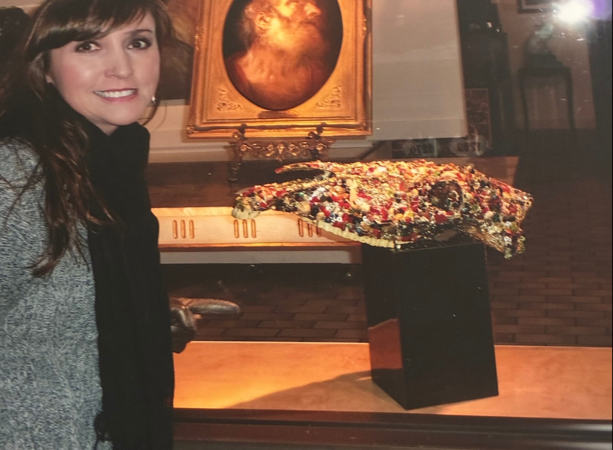 Smiling woman with dark hair wearing a gray coat, beside a colorful flower arrangement at an indoor altar. Note: concise description of subject, setting, and key elements.