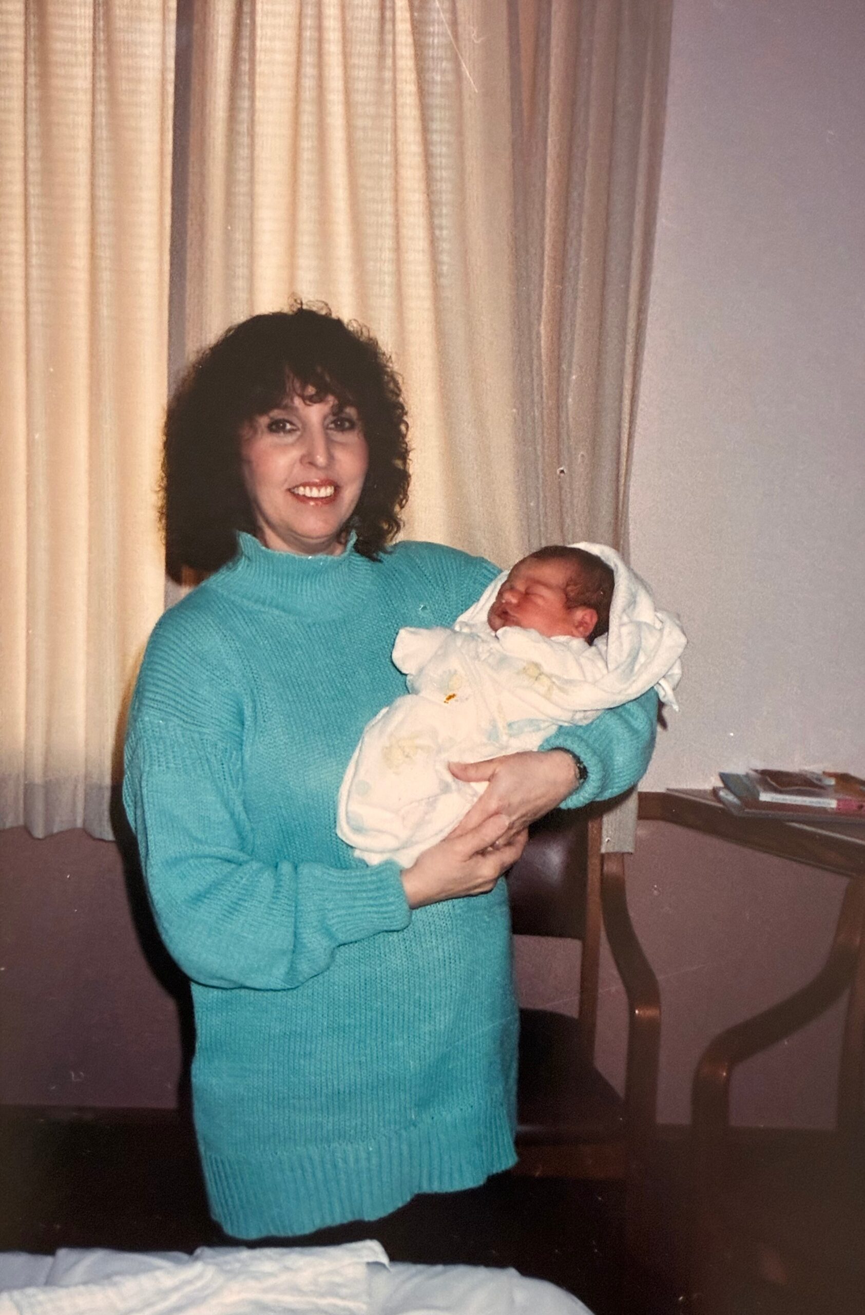 A smiling woman in a turquoise sweater holds a newborn baby wrapped in a white blanket indoors by curtains.