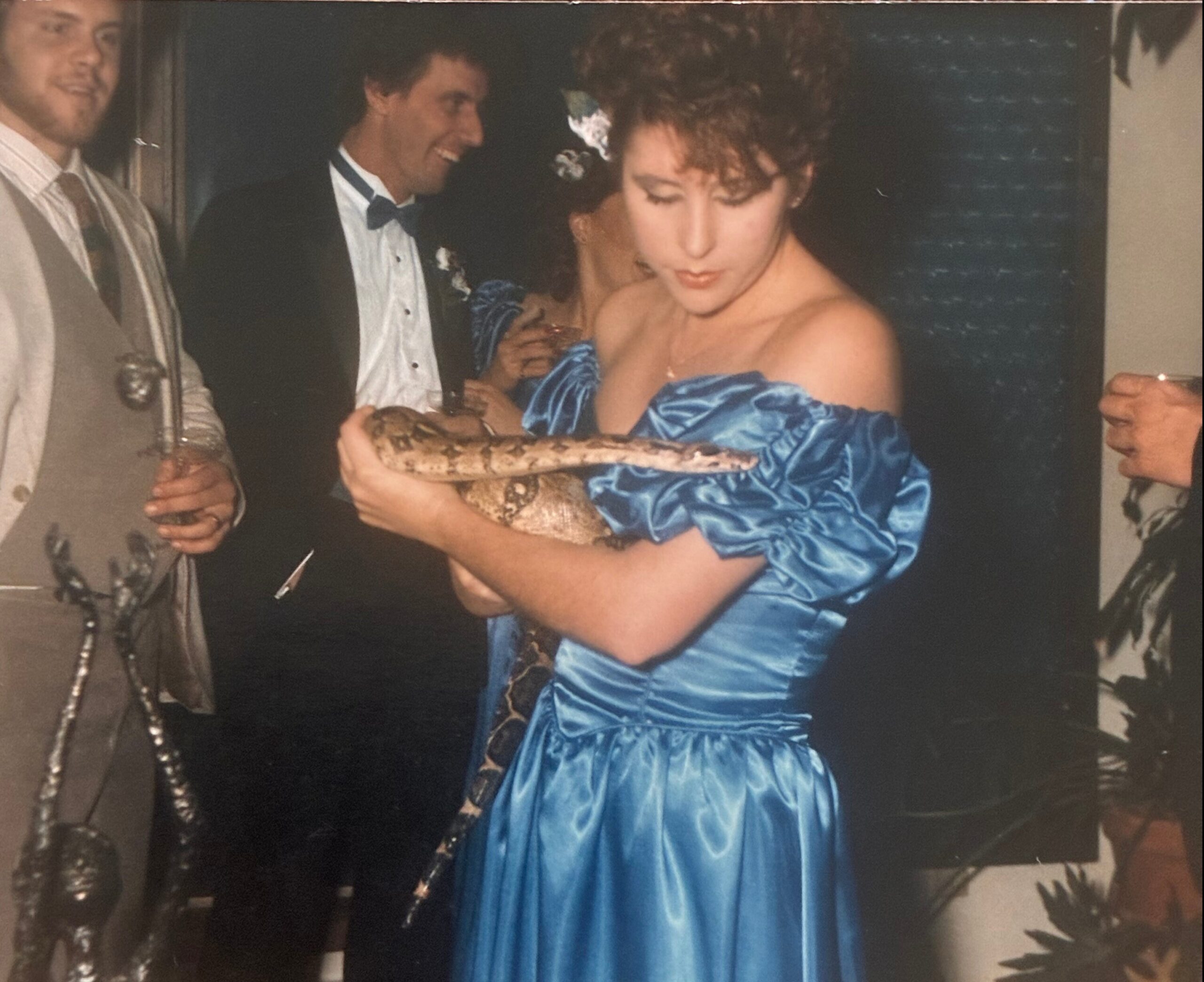 Woman in a blue satin off-shoulder gown holding a large snake at a formal party, with men in tuxedos nearby.