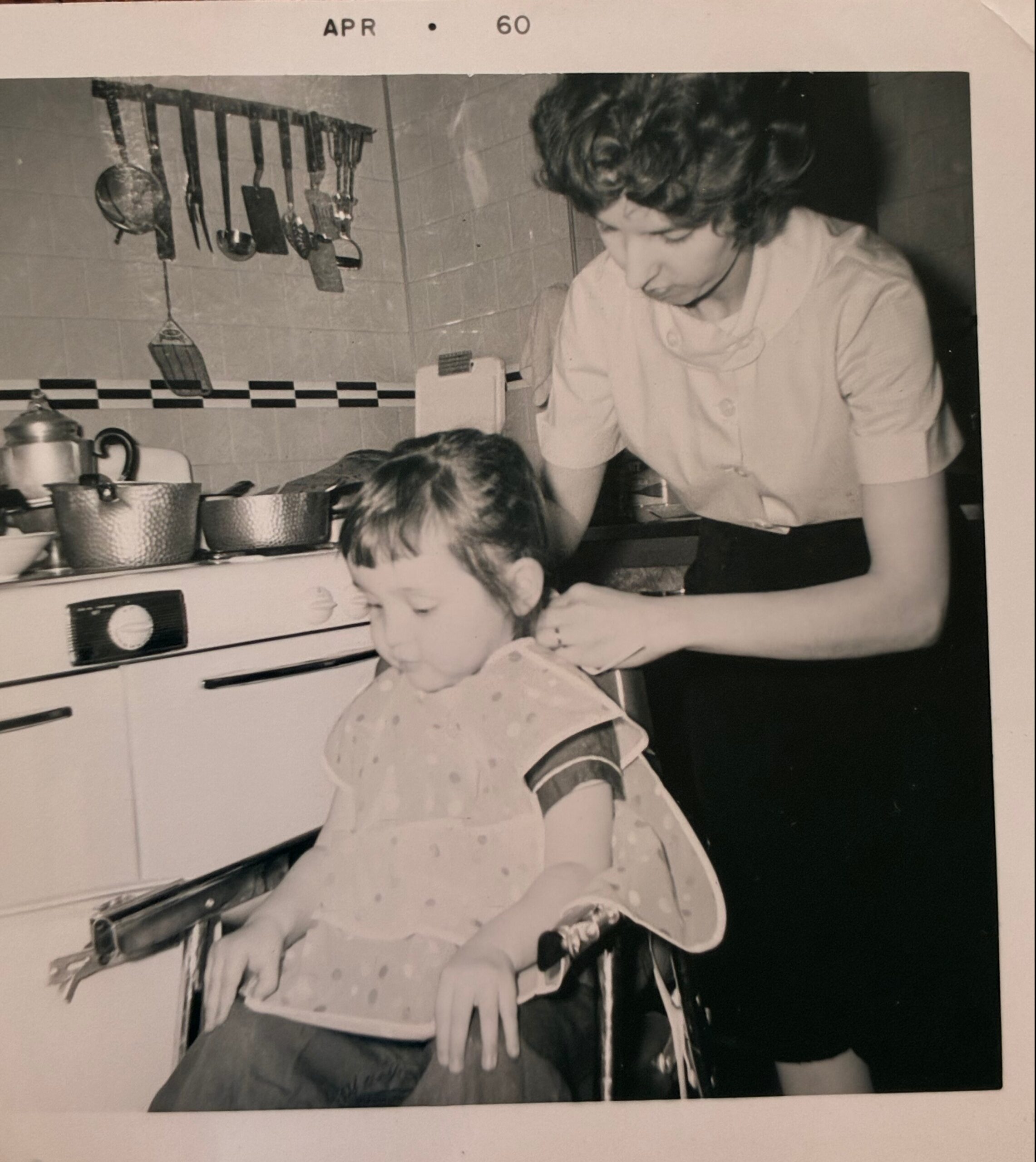Woman leaning over a child at a kitchen counter as they prepare food, with pots and utensils hanging on a tiled wall in a vintage kitchen photo.