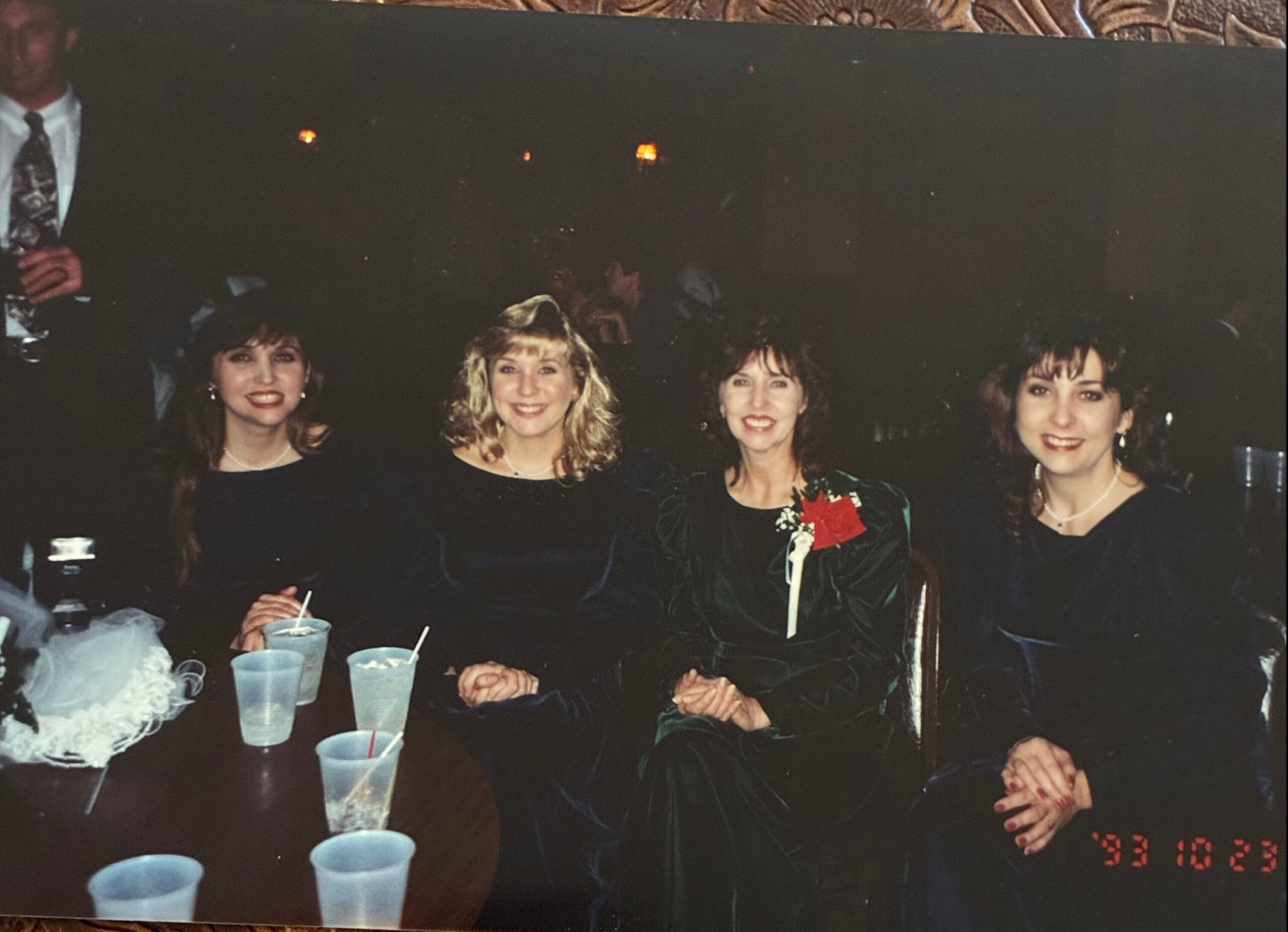 Four women in formal dresses sit around a table at a party, smiling for the photo with blue cups in front of them. A date stamp reads 93 10 23 in the bottom corner.