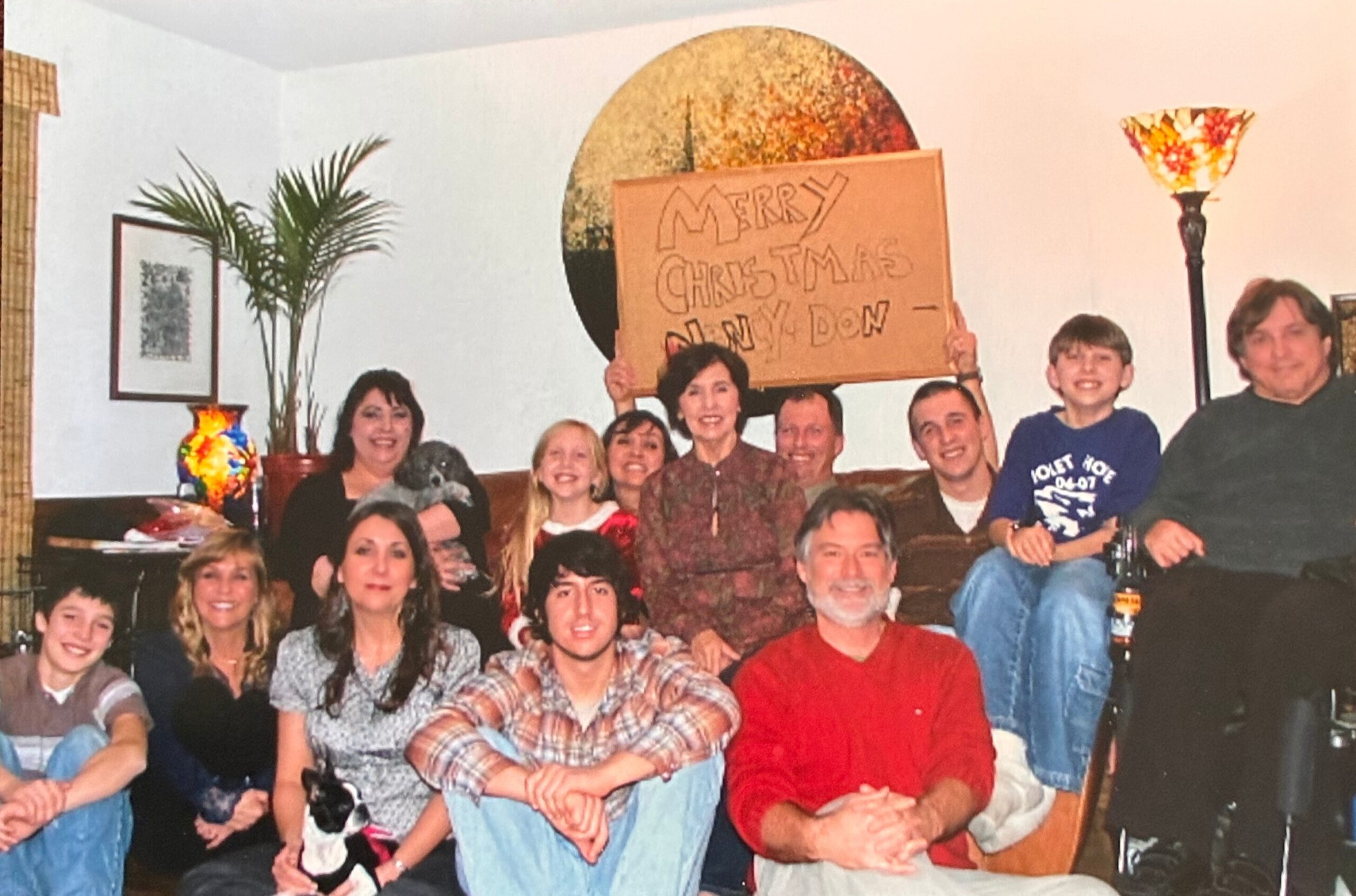 Family and friends posing for a Christmas photo indoors, with a cardboard sign reading 'Merry Christmas'.