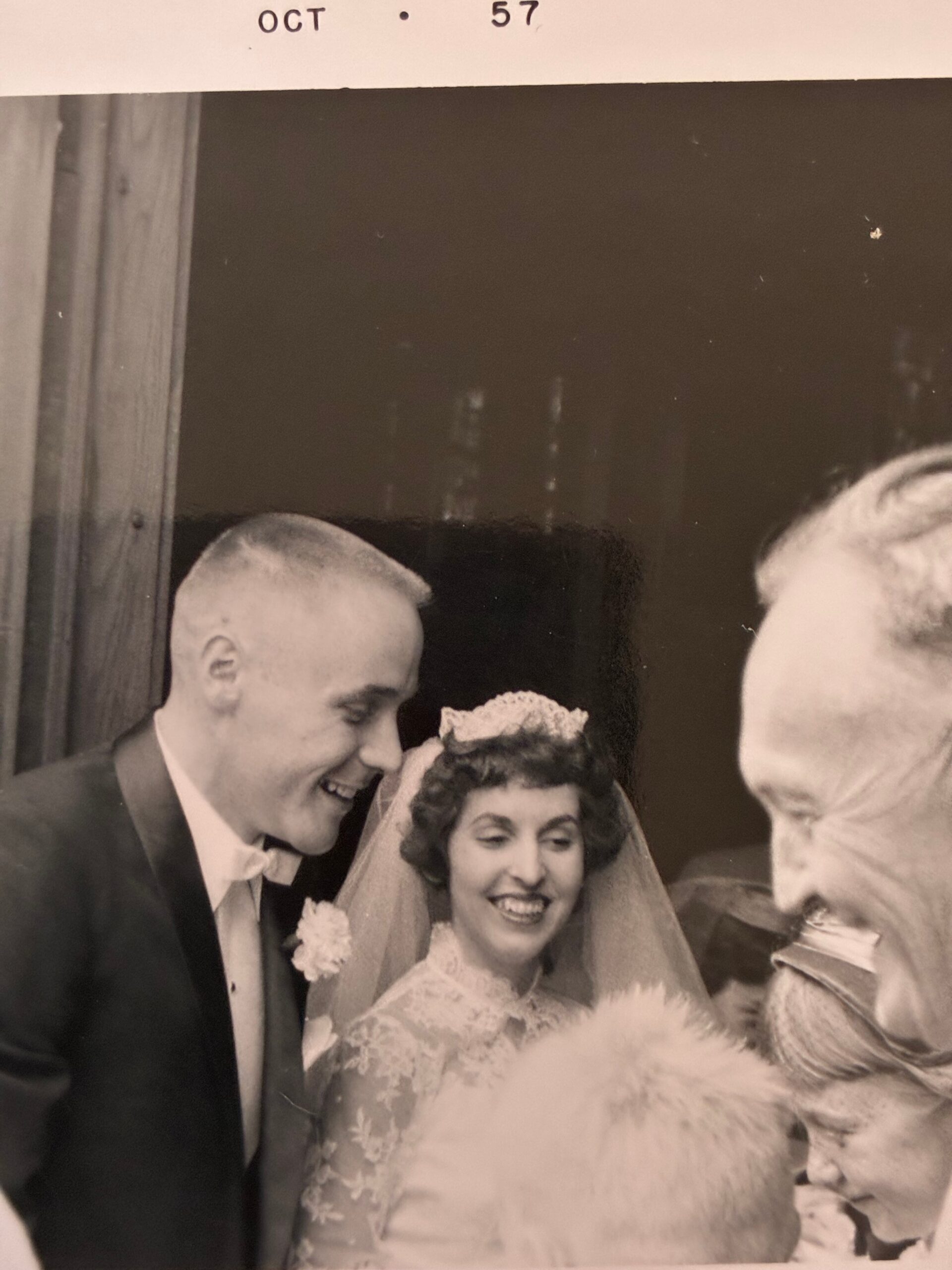 Black-and-white wedding photo: smiling bride in lace gown and veil beside groom in tux, with an October 1957 stamp above them.