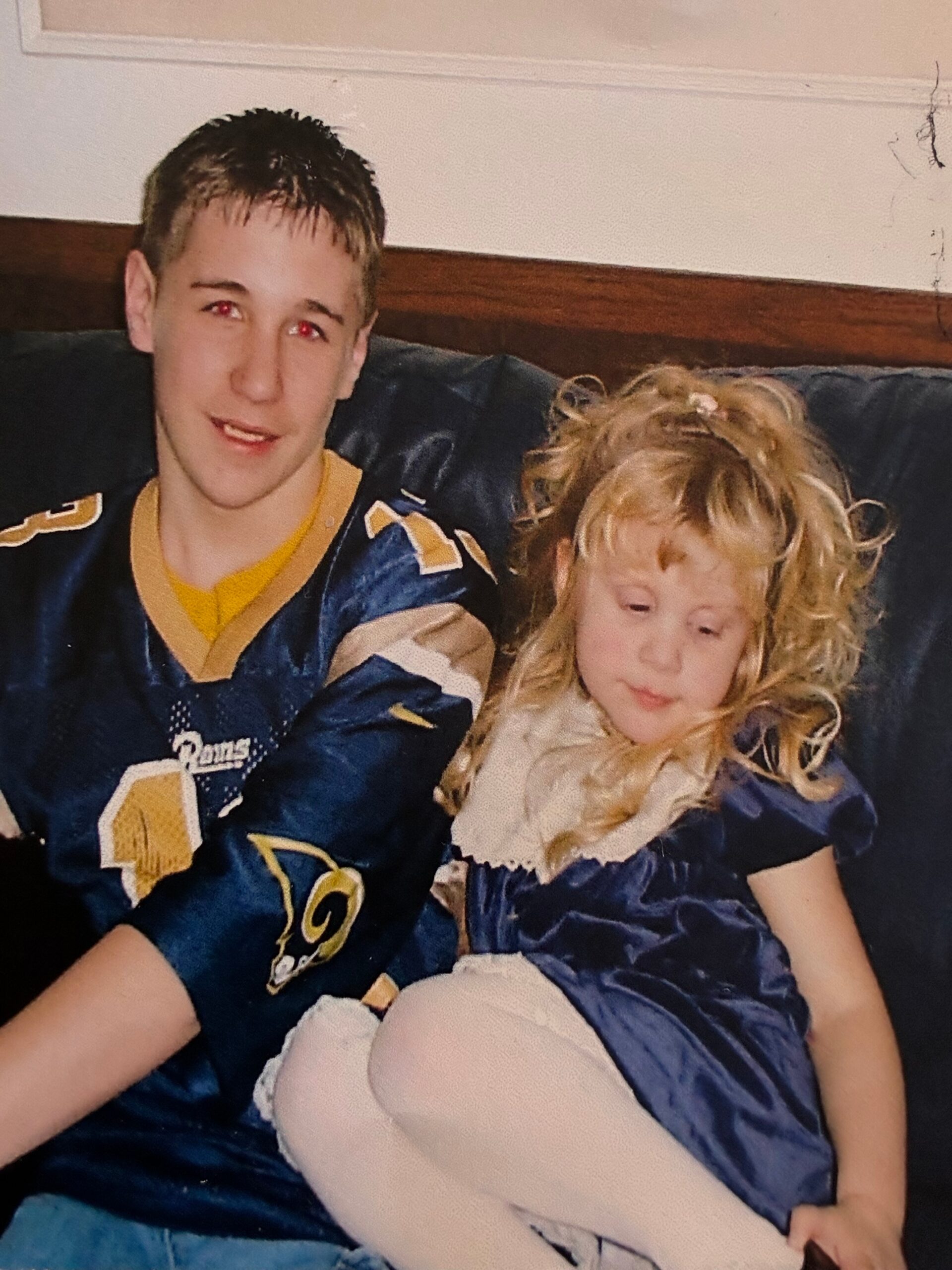 Teen boy in a blue and gold football jersey sitting on a sofa with a young girl in a blue dress on the same couch.