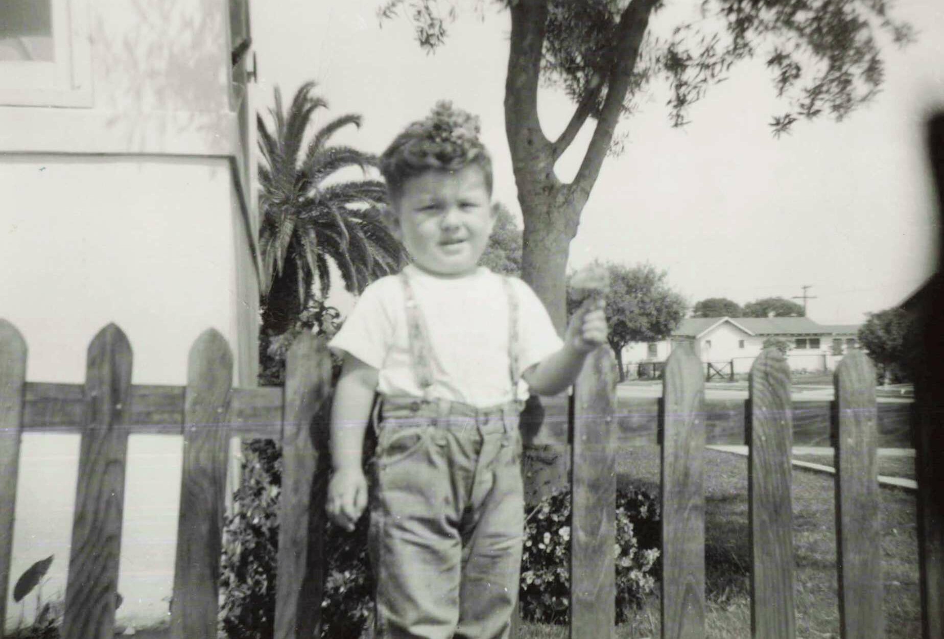 Young boy standing behind a wooden picket fence in a residential yard, wearing suspenders and a white shirt, smiling at the camera.