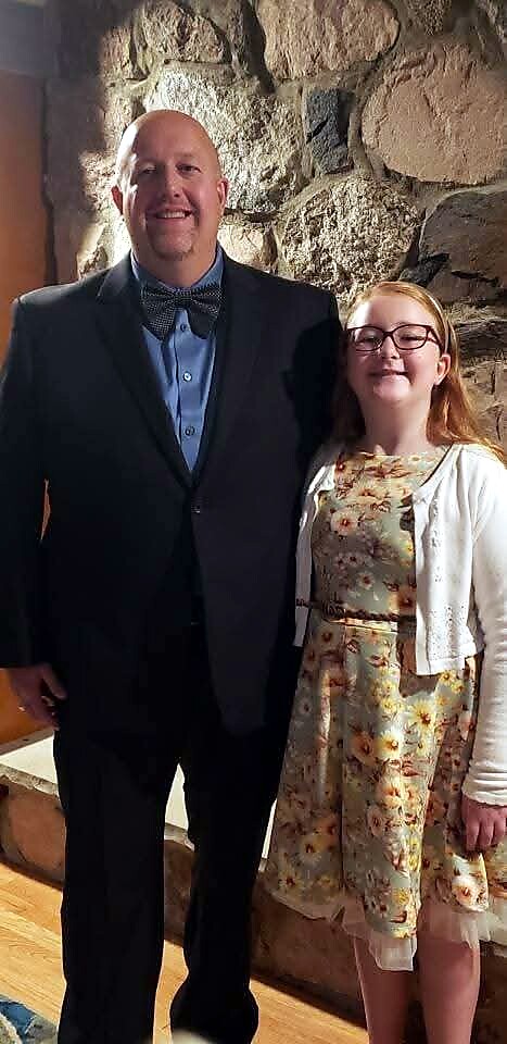 Two people posing for a photo: a tall man in a dark suit and bow tie beside a girl in a floral dress with glasses, against a stone wall indoors.