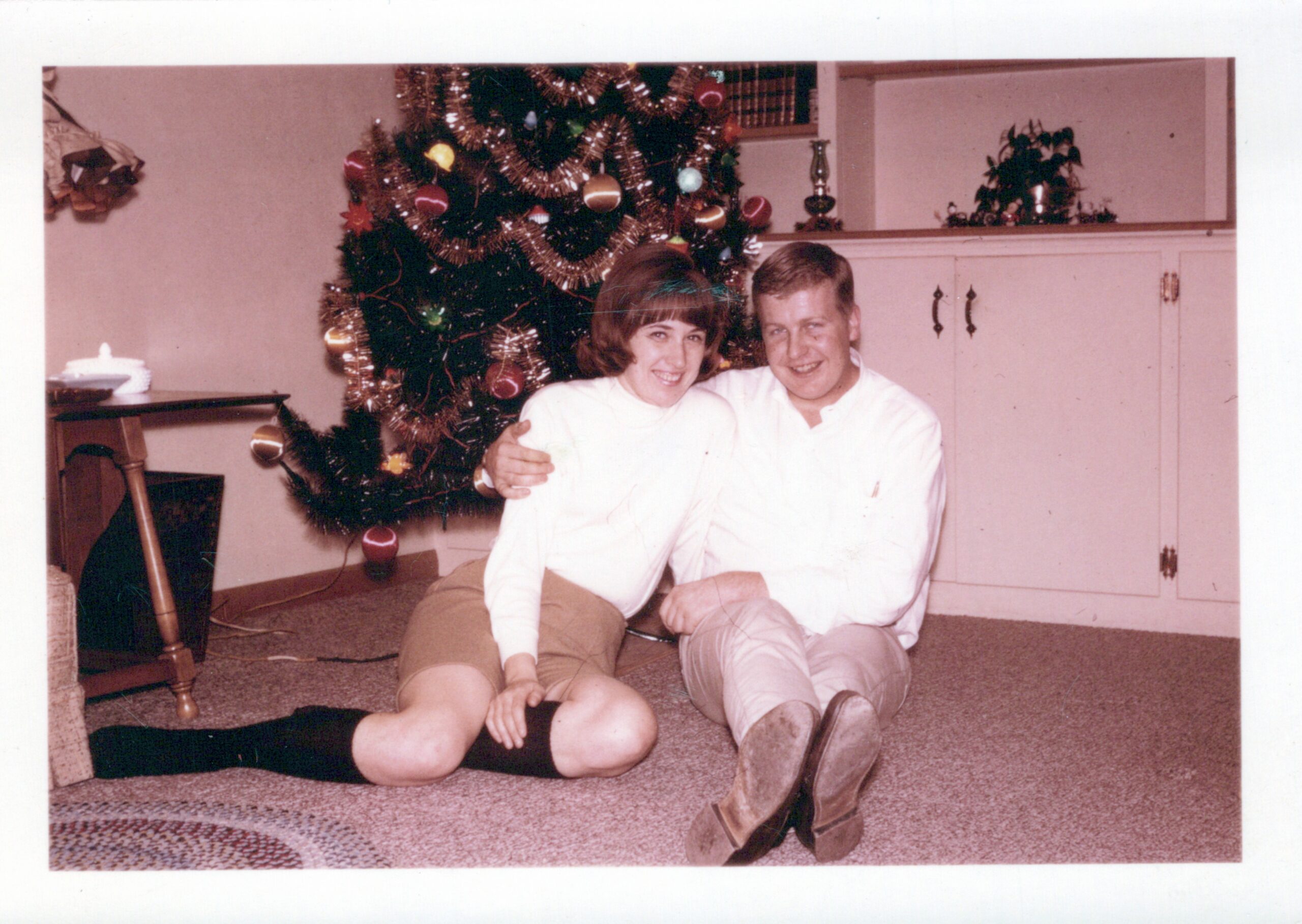 Smiling couple sitting on the carpet in front of a decorated Christmas tree in a living room.