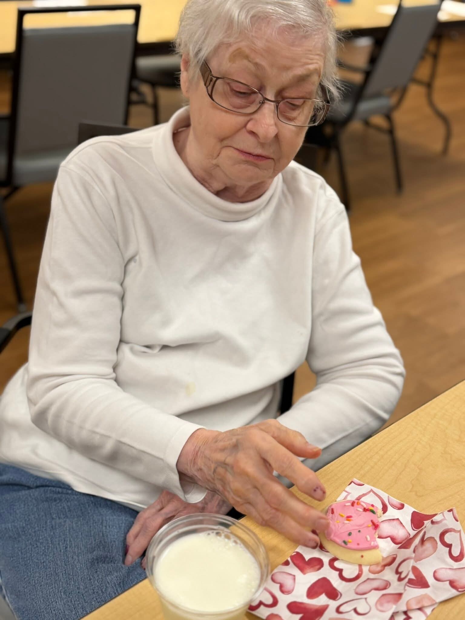 Elderly woman with glasses in a white sweater sits at a wooden table, about to eat a pink frosted donut with sprinkles beside a glass of milk on a heart-patterned napkin.