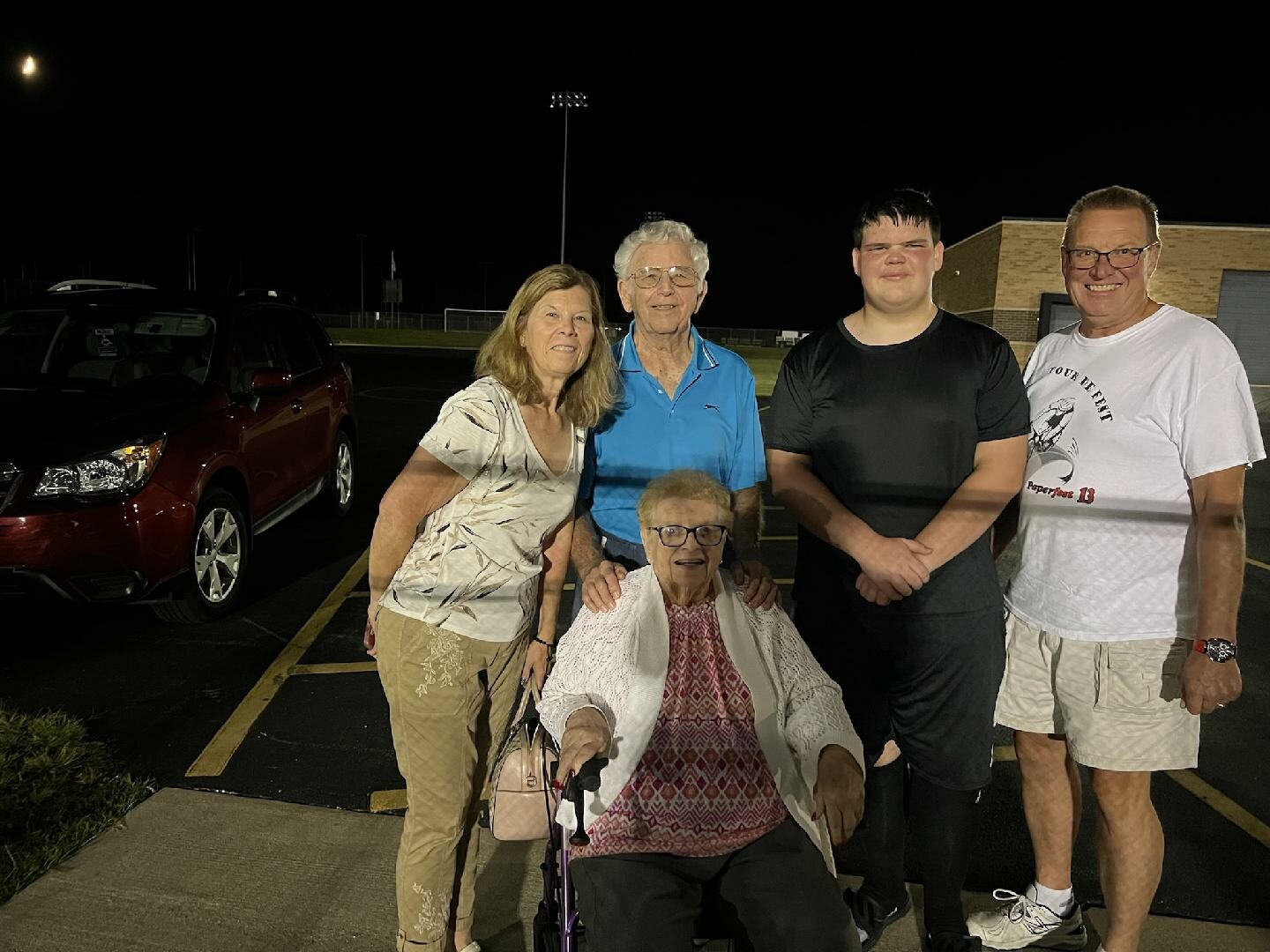 Five people pose in a dim parking lot at night; an elderly woman in a wheelchair sits in front while four others stand behind her near a red SUV.